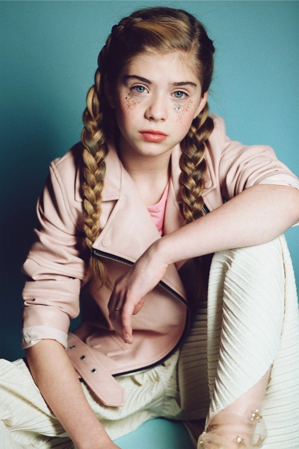 Young girl with braided hair wearing a pink jacket and light-colored pants, seated against a blue background.