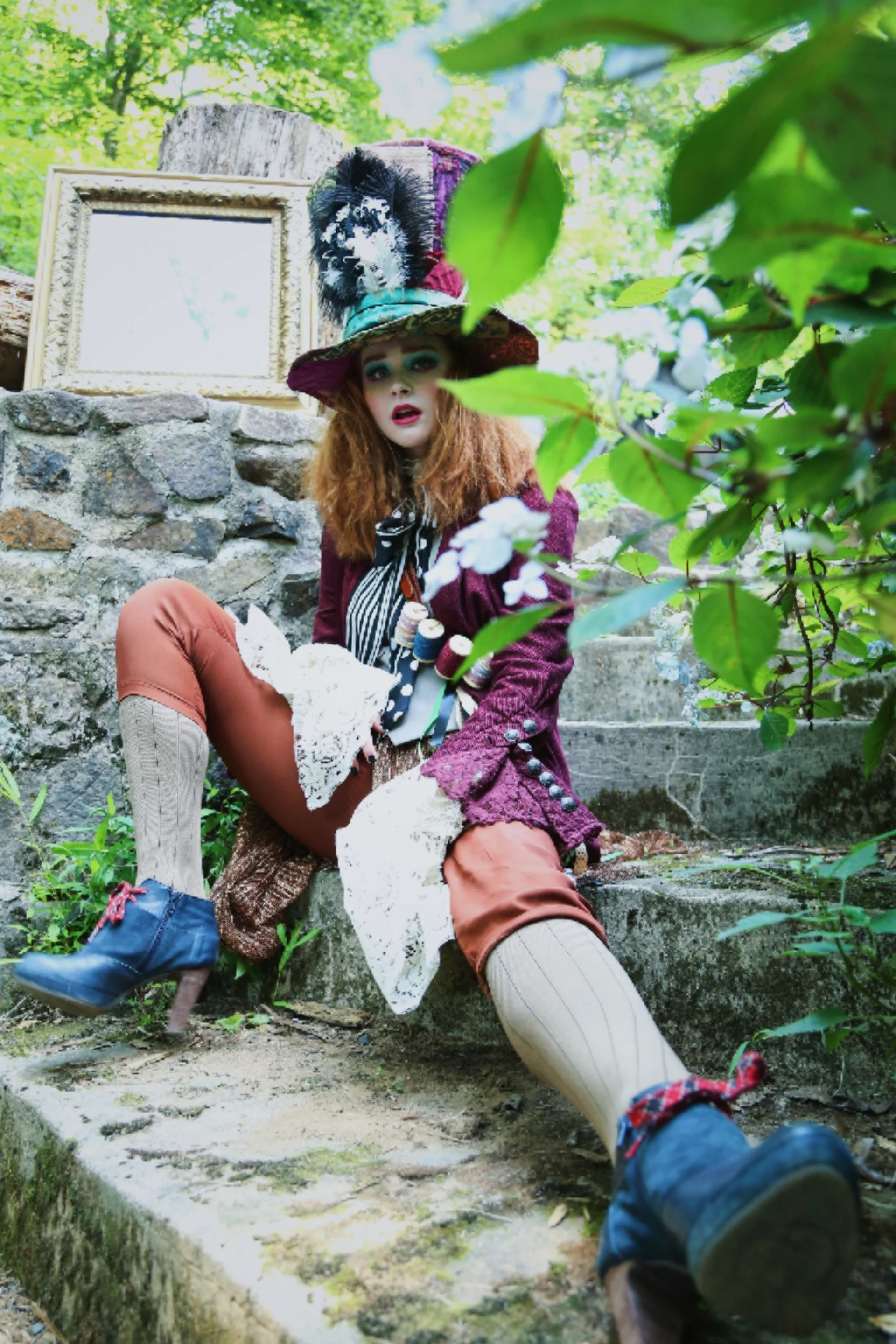 Person in eccentric costume resembling Mad Hatter, with colorful hat and vintage clothing, sitting on outdoor stone steps surrounded by greenery and a framed mirror.