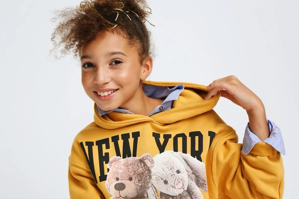 Smiling child in a yellow hoodie with 'New York' text and teddy bear prints.