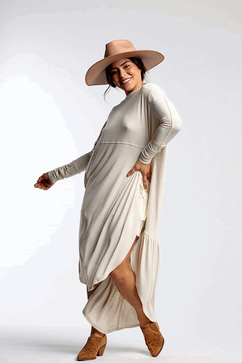 Woman wearing a cream maxi dress, tan hat, and tan ankle boots, posing playfully against a plain background.