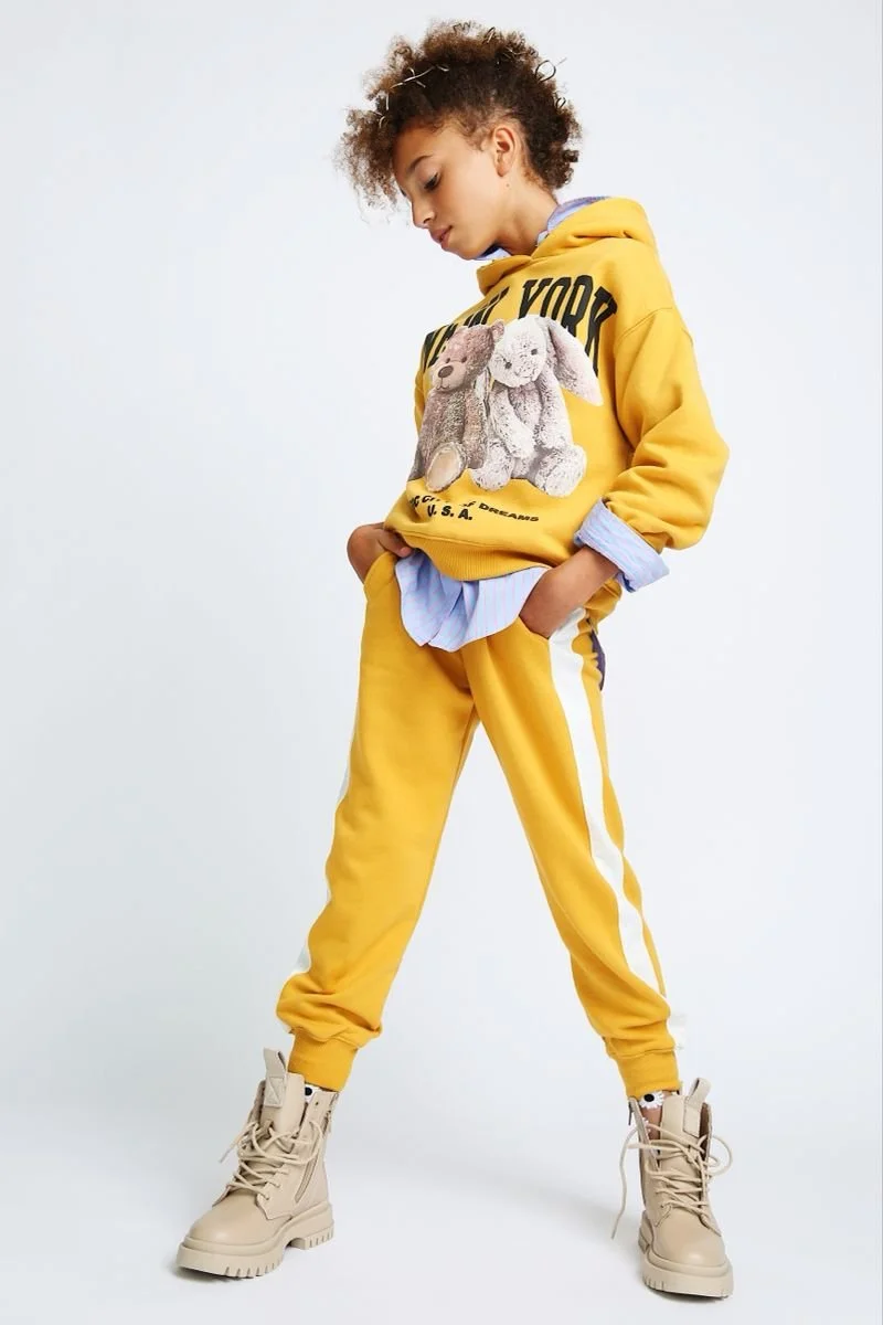 Child in yellow hoodie and sweatpants with teddy bear print, wearing beige boots, standing confidently.