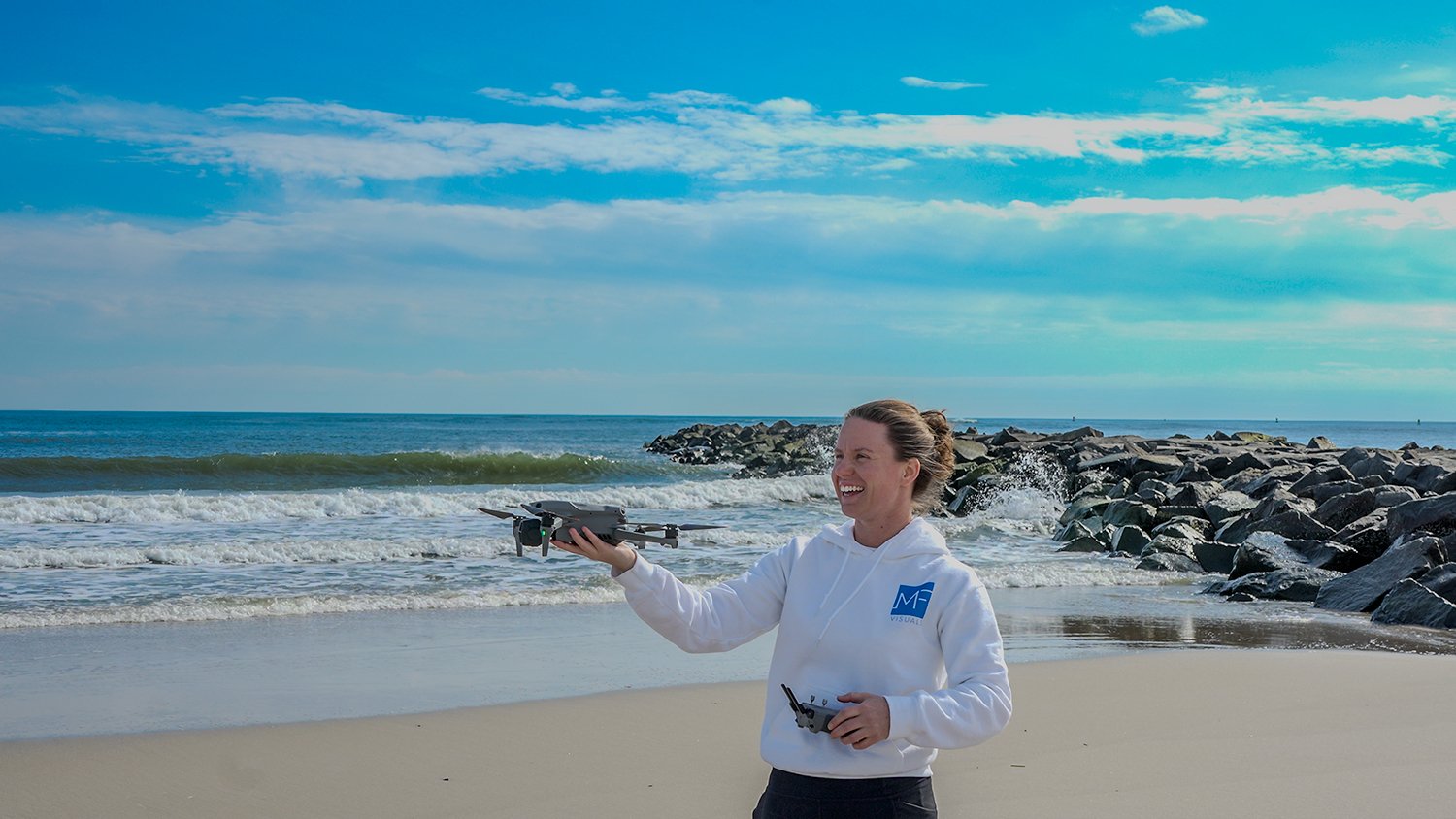Woman in white hoodie smiling while flying a drone on beach with rocks and ocean in background.
