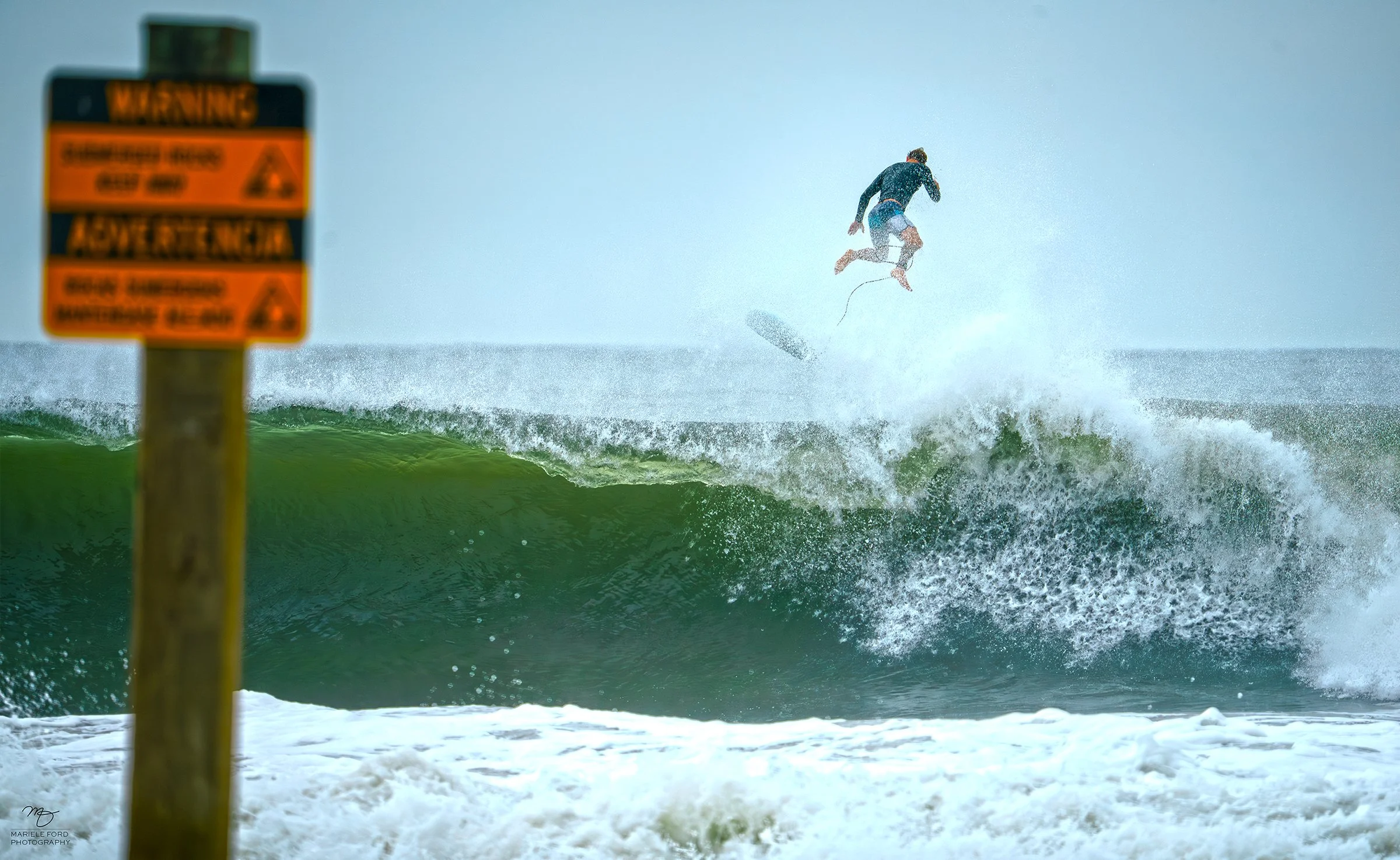 A person surfing on a large green wave near the shore, with a warning sign in the foreground.