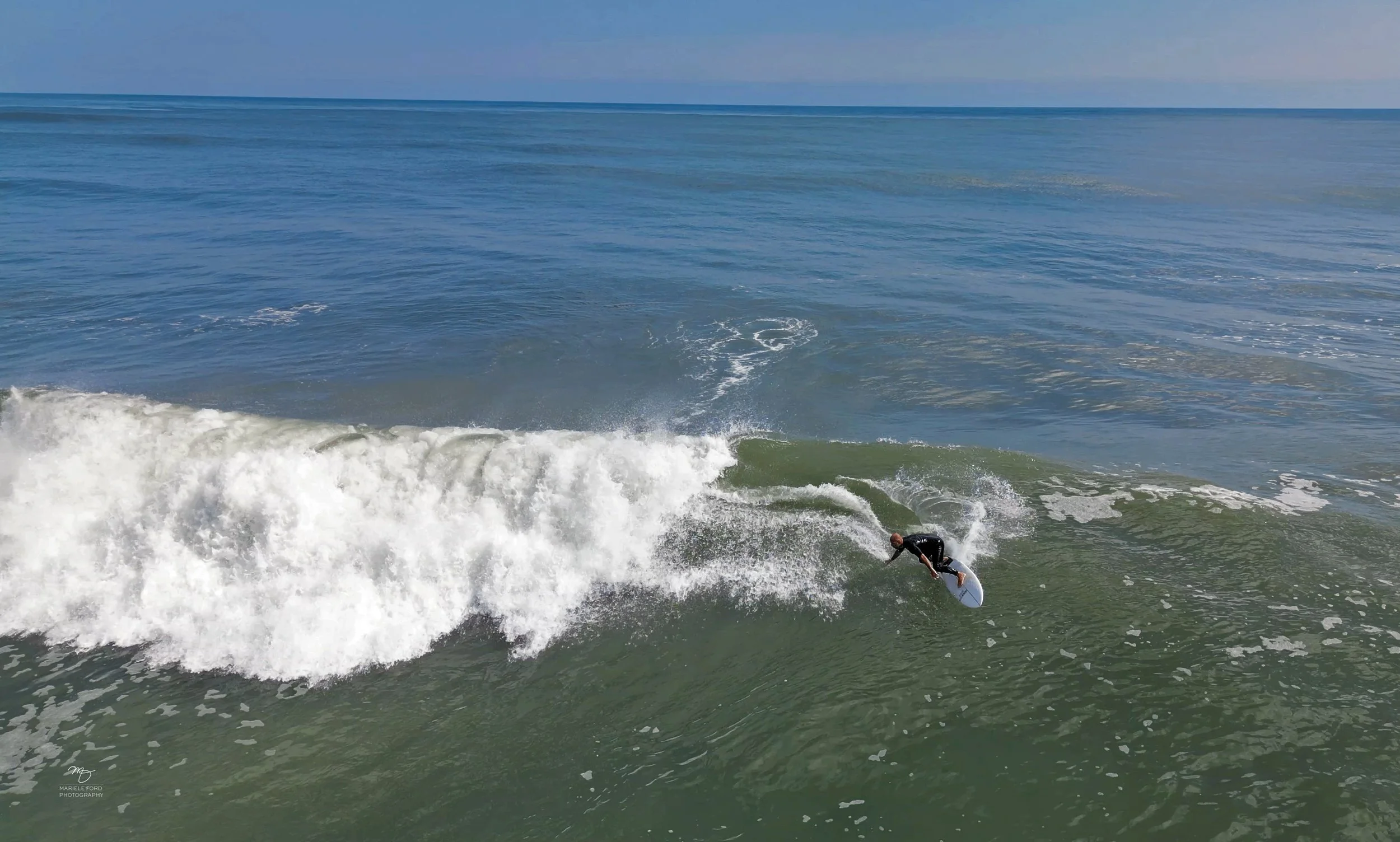 A person surfing on a wave in the ocean, wearing a wetsuit, with open sky in the background.