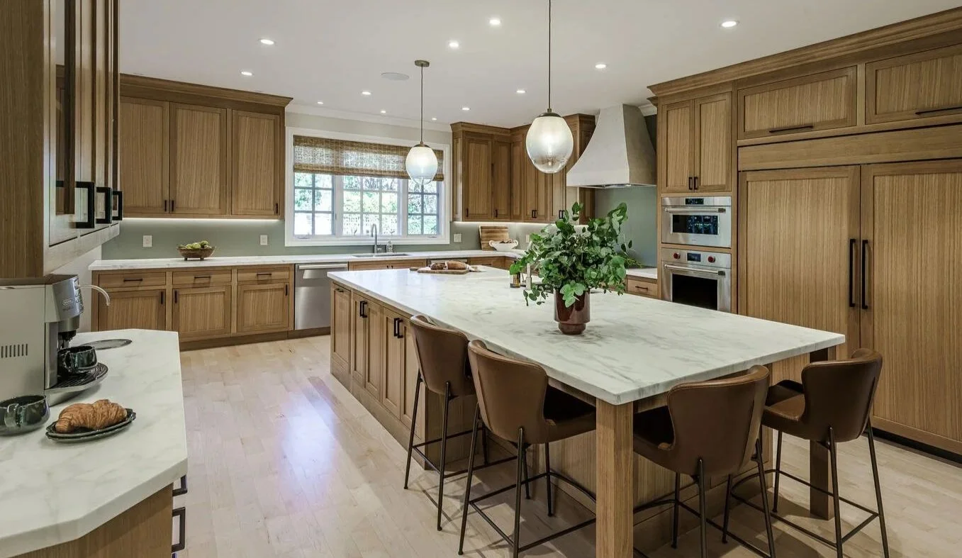 Modern kitchen with wooden cabinets, a marble island with seating, pendant lighting, a window with woven shades, and stainless steel appliances.
