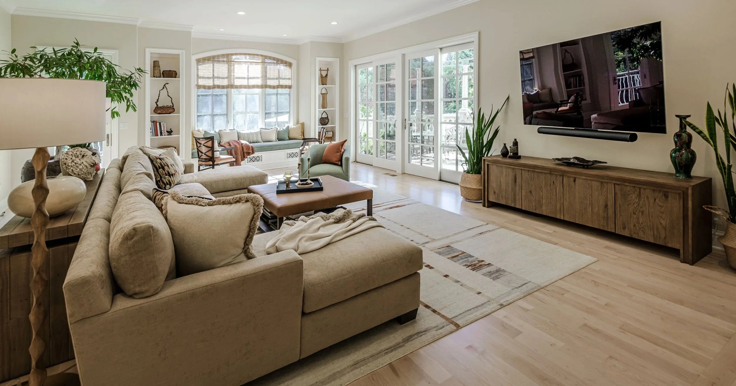 Bright living room with beige sectional sofa, wooden coffee table, large area rug, built-in window seat with cushions, and a flat-screen TV mounted on the wall, with glass doors leading to a patio.