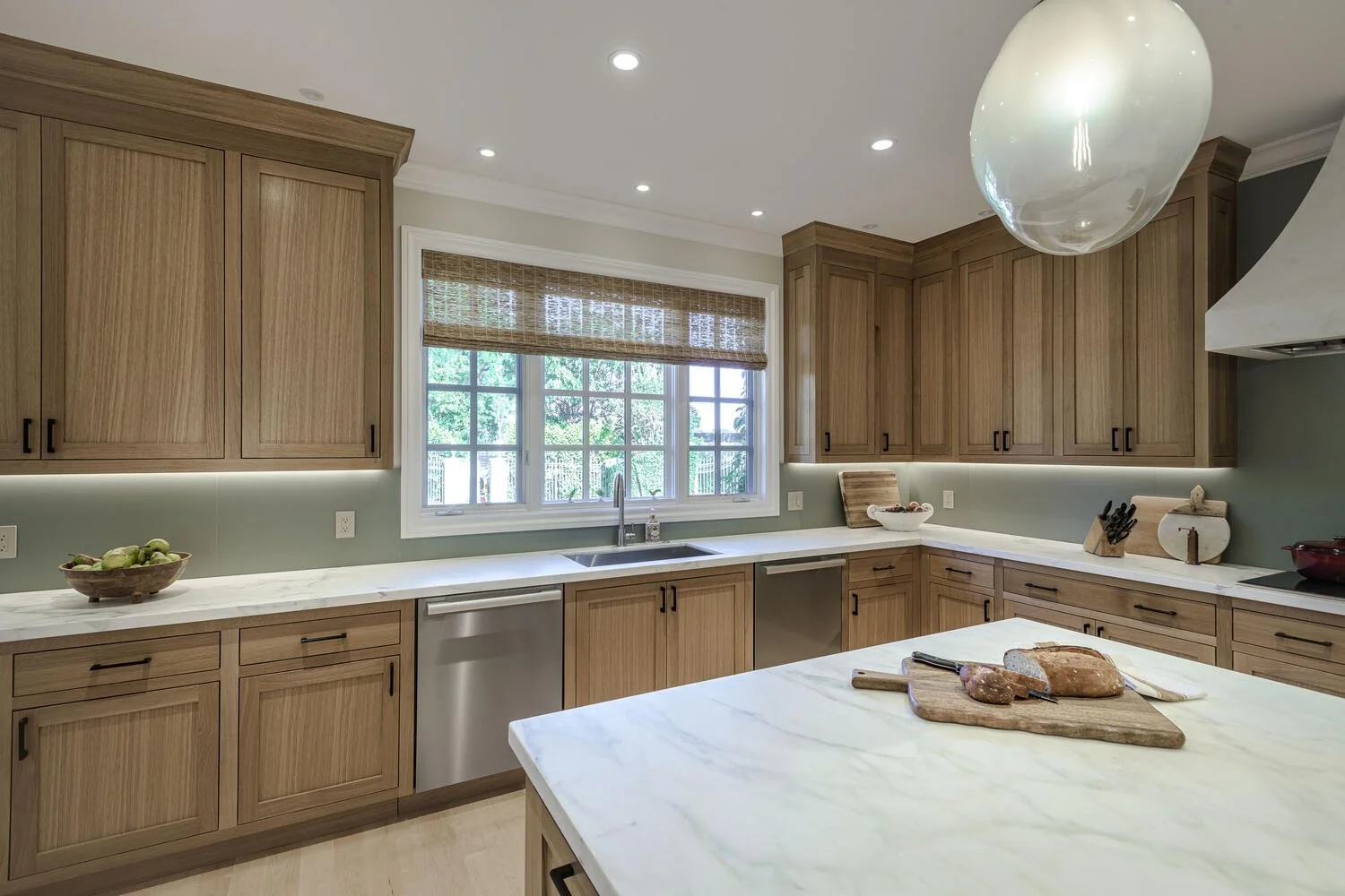 Modern kitchen with wooden cabinets, white marble countertops, a window with bamboo blinds, and a kitchen island with bread and knives.
