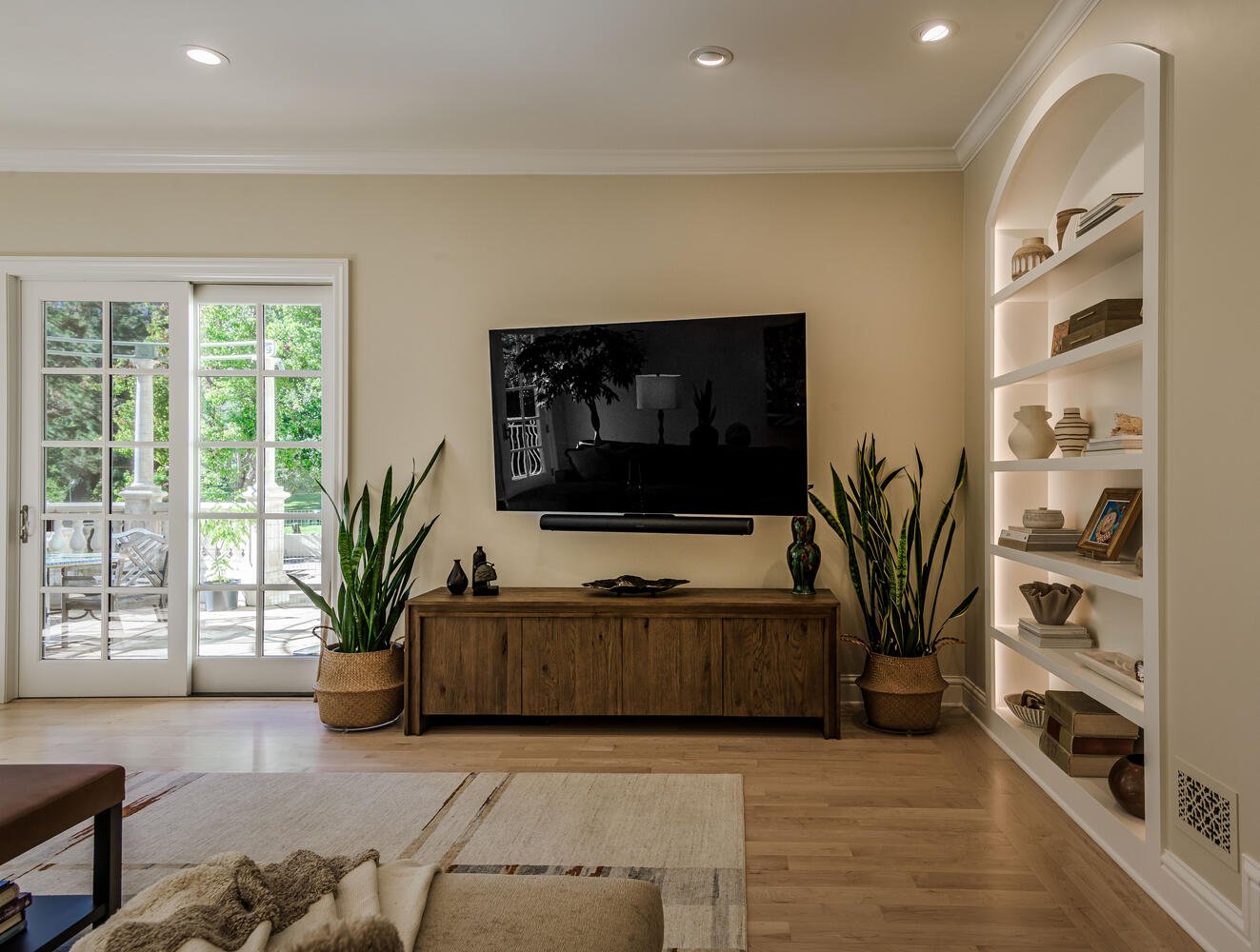 Living room with sliding glass doors leading to a patio, wall-mounted TV, wooden console table with decorative items, large potted plants on either side, built-in bookshelf with decorative objects, light-colored walls and hardwood floor.