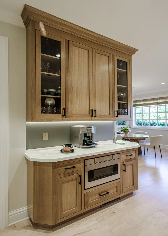 Wooden kitchen cabinetry with glass-front upper cabinets, white marble countertop with a coffee maker, and a small cookie on a plate. Open living area with large windows in the background.