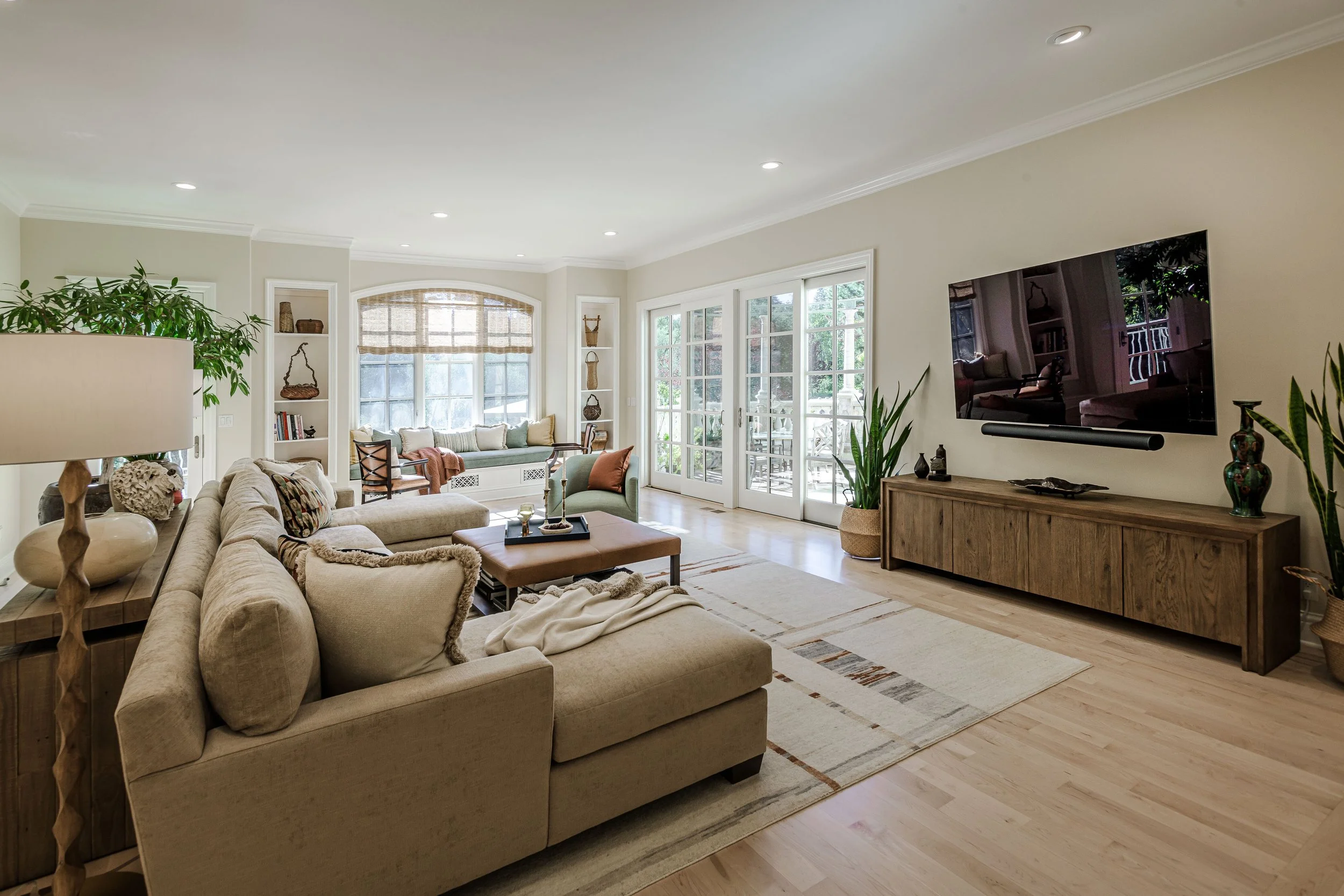 Bright living room with beige sectional sofa, wooden coffee table, large area rug, built-in window seat with cushions, and a flat-screen TV mounted on the wall, with glass doors leading to a patio.