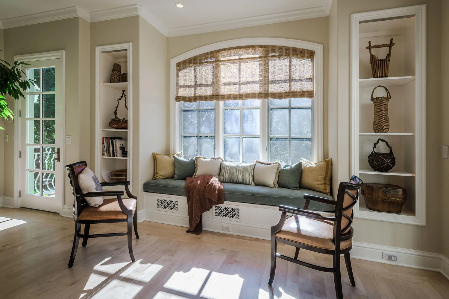 Cozy living room nook with a large window seat decorated with multiple cushions, flanked by built-in white shelves filled with decorative baskets, books, and decor items. A brown blanket is draped over the seat. Two wooden chairs with cushioned seats