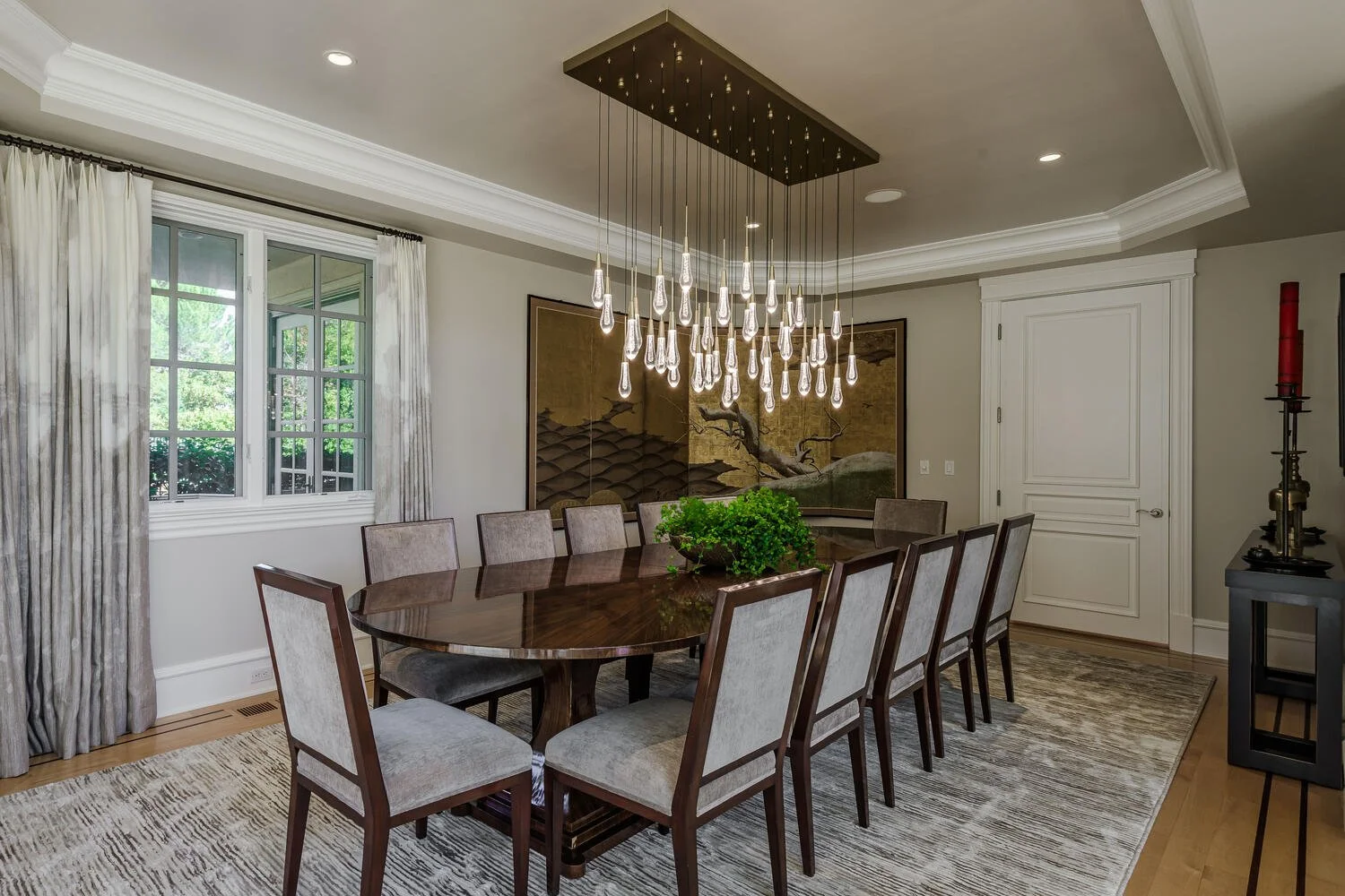 Elegant dining room with a large wooden table, 10 upholstered chairs, modern chandelier, large window with white curtains, Japanese-style folding screen, and artwork on the walls.