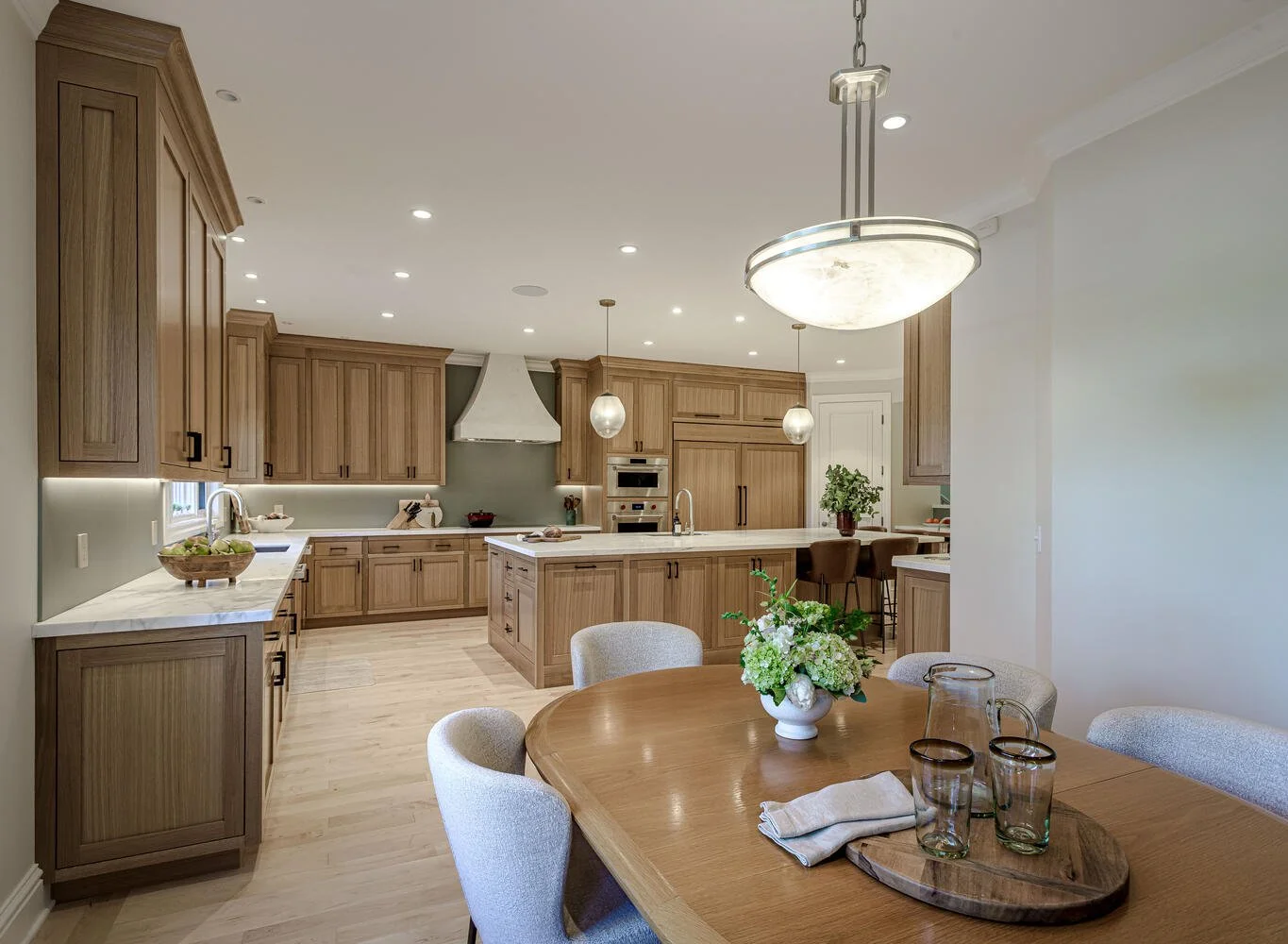 Open kitchen with wooden cabinets, a central island, marble countertops, and pendant lighting, adjacent to a dining area with a wood table, vase with white flowers, and drinking glasses.