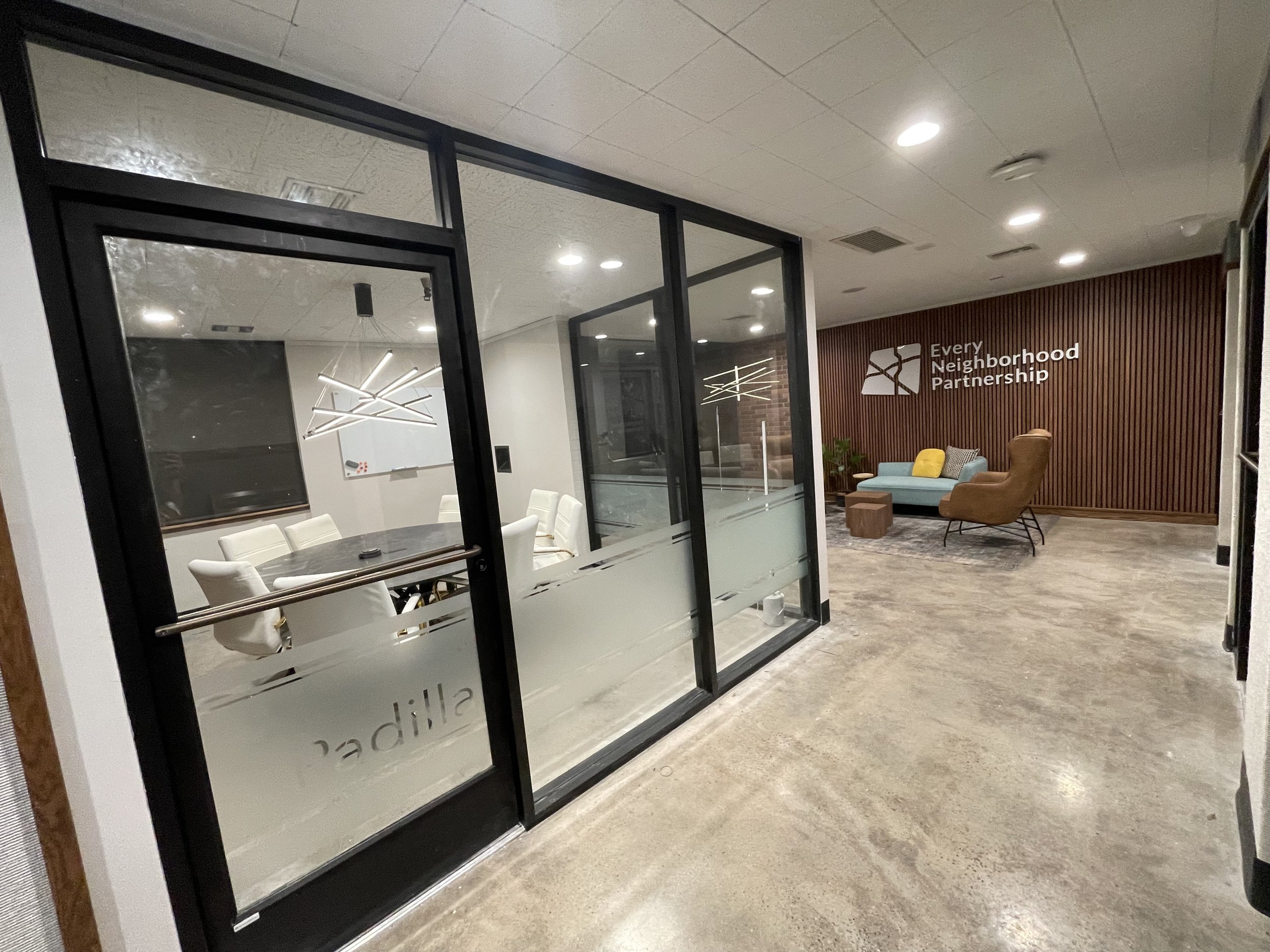 Modern office or community space with glass-walled conference room on the left featuring a large round table and eight white chairs, and a cozy lounge area on the right with a blue sofa, a yellow pillow, a brown armchair, and a small wooden coffee table against a wood-paneled wall with the logo and text "Every Neighborhood Partnership."