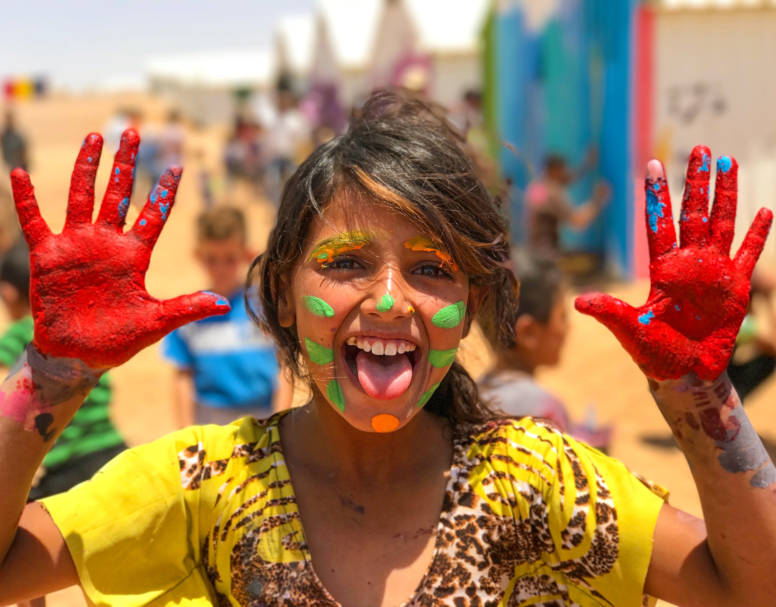 A person with colorful face paint and hands covered in red paint, smiling and holding up their hands outdoors, with blurred people and tents in the background.