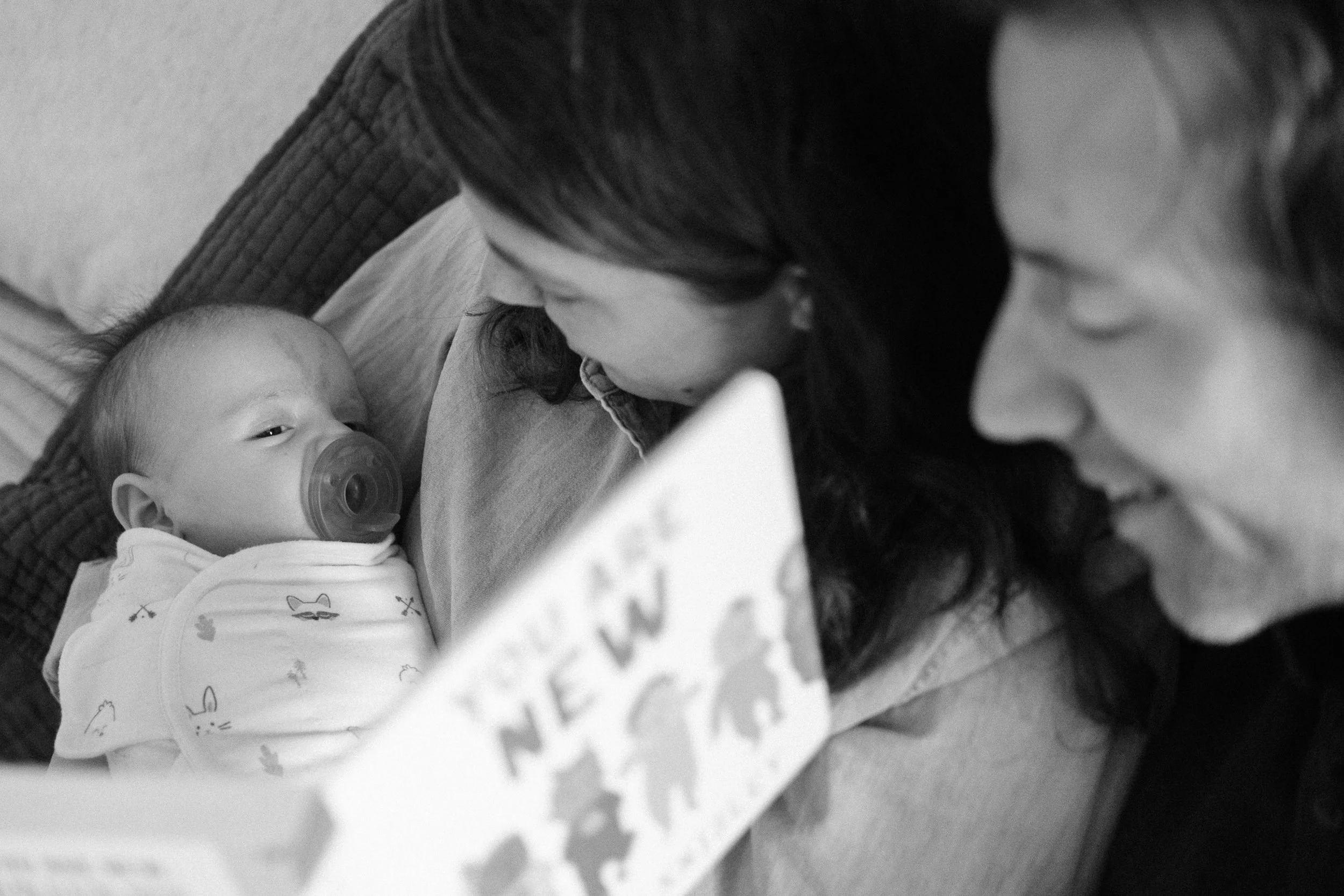 An infant with a pacifier lying on a woman's chest, while another woman looks on, reading a book titled 'You Are New'.