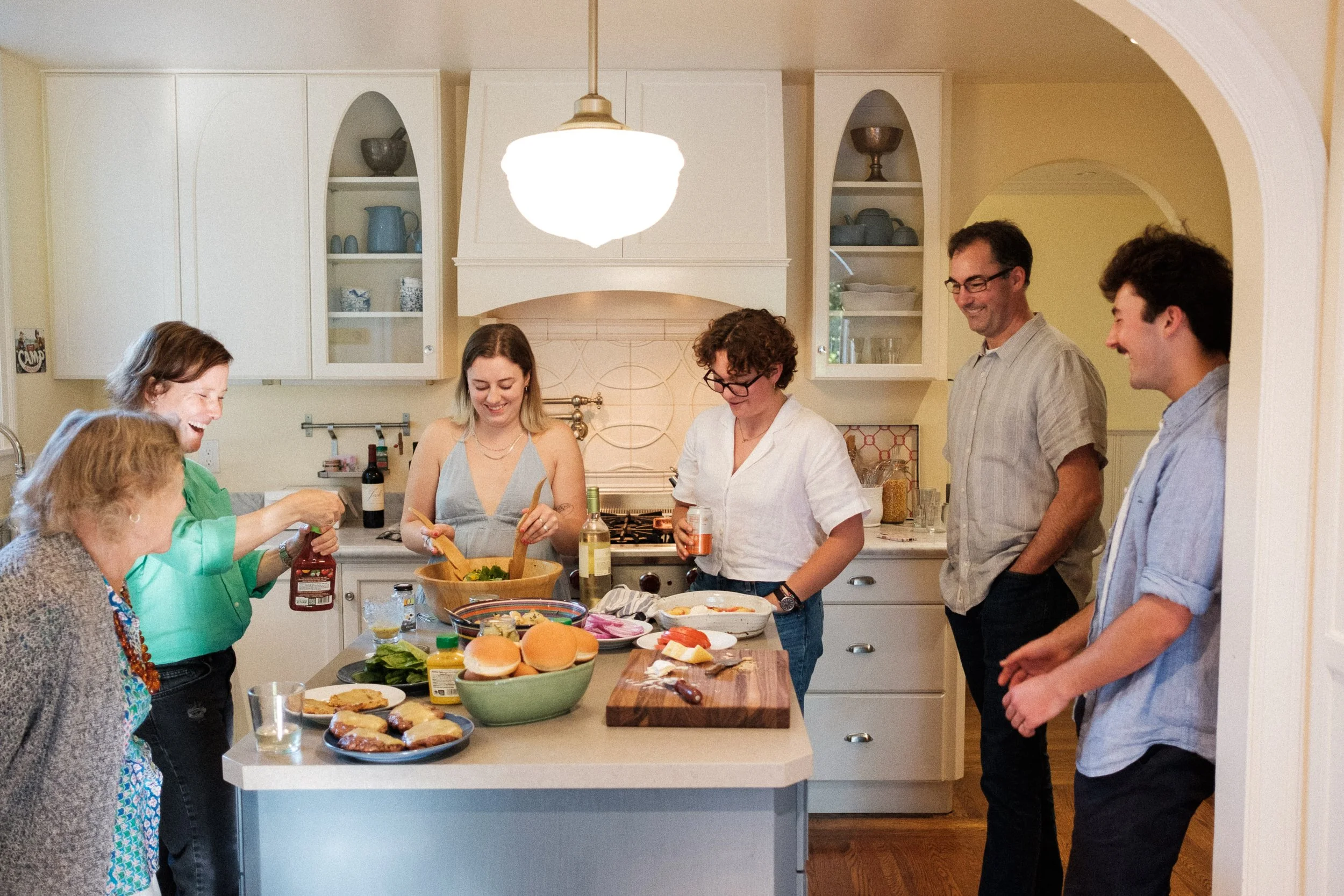 Group of six people in a kitchen preparing and enjoying a meal together, smiling and laughing.