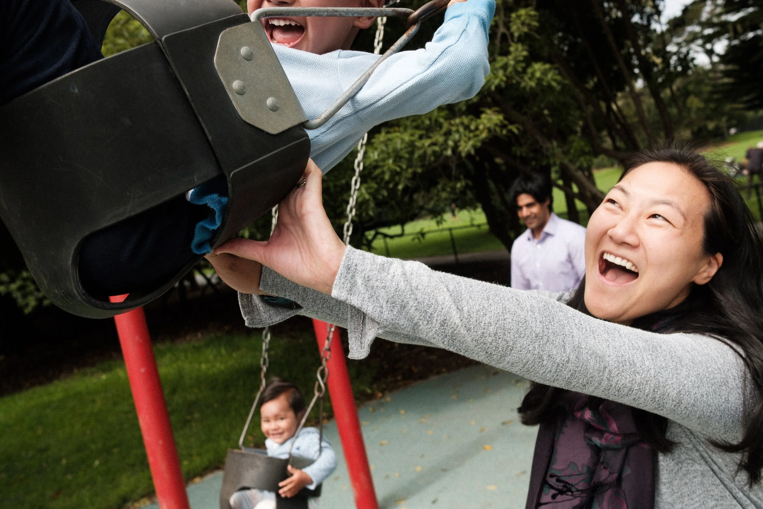 A woman joyful while helping a young boy in a swing set at a park, with a girl smiling in a swing in the background.