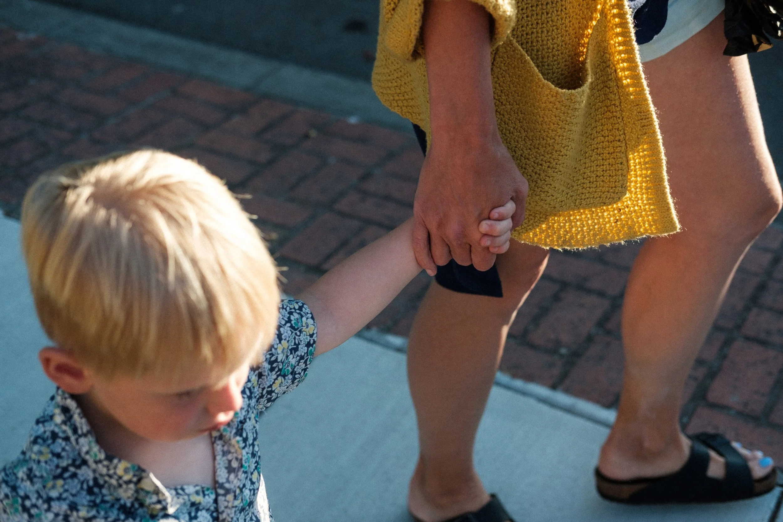A young boy with blonde hair and a floral shirt holding hands with an adult woman wearing a yellow cardigan and black sandals, walking on a sidewalk.