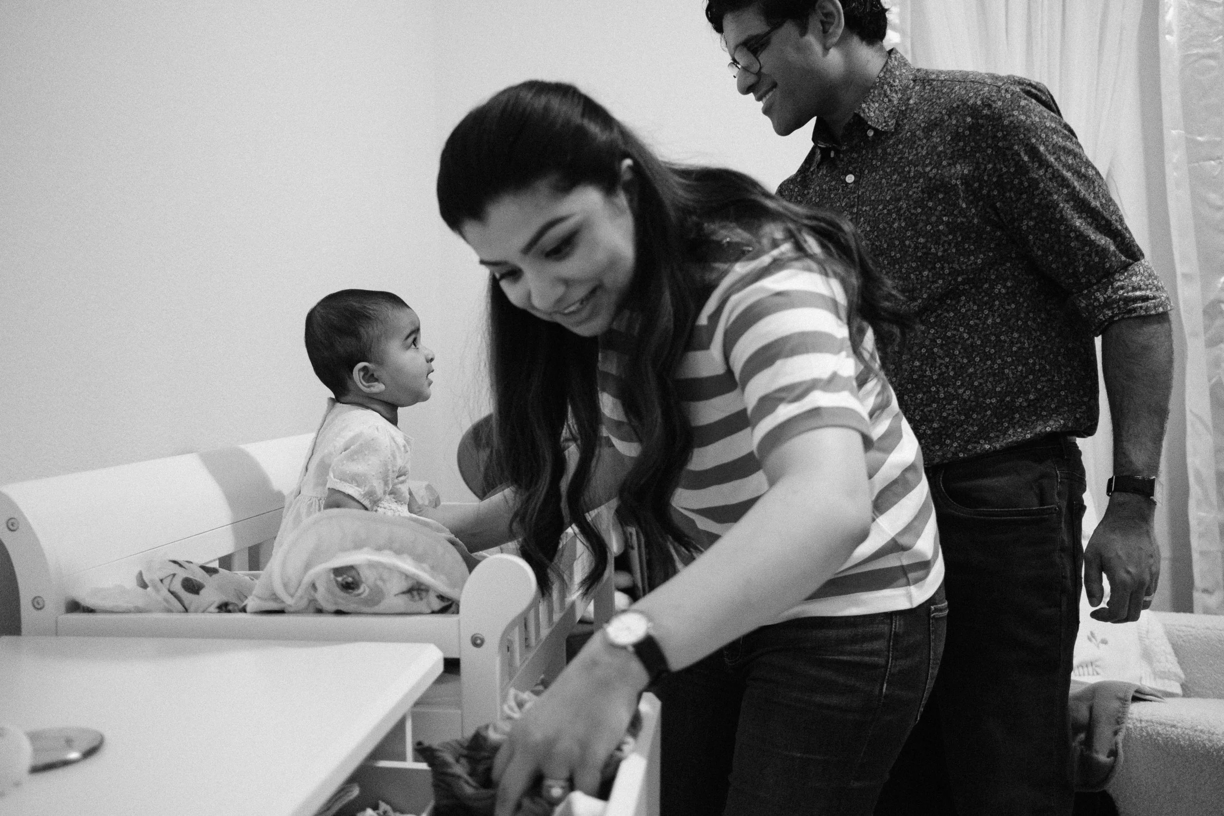 People in a room, a woman leaning over a table, a man smiling standing behind her, a small child sitting on a bed, a teddy bear and blanket on the bed, in a cozy room with curtains.