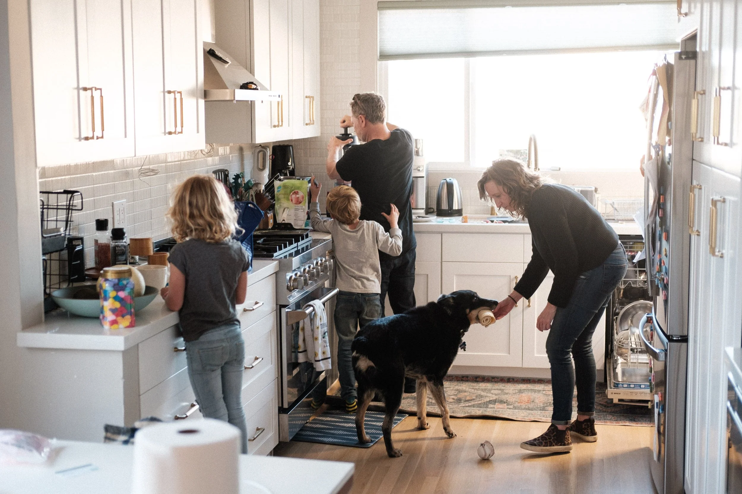 A family in a bright, modern kitchen. A woman is feeding a dog with a chew toy in its mouth. Two children are preparing food at the counter, and a man is near the window, focused on an activity.