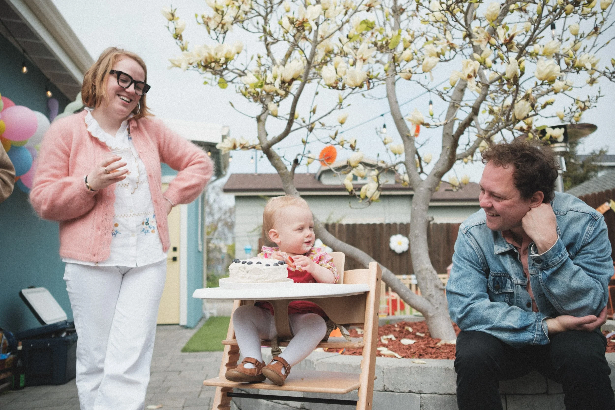 A family celebrating a young child's birthday outdoors, with a woman standing, a man sitting, and a toddler in a high chair with a birthday cake, under yellow flowering trees.