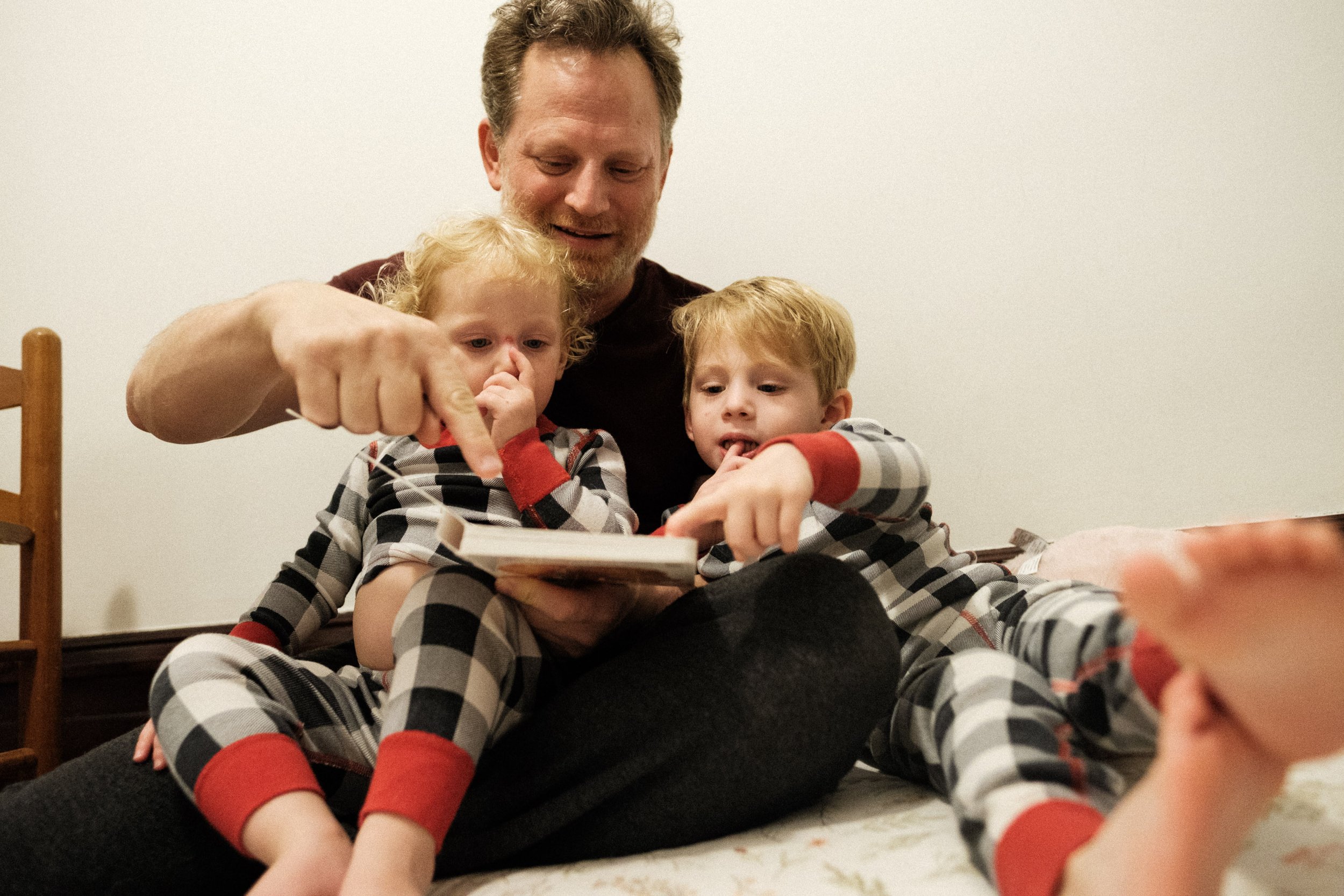 A father and two young children in matching pajamas sitting on a bed, looking at a book together.