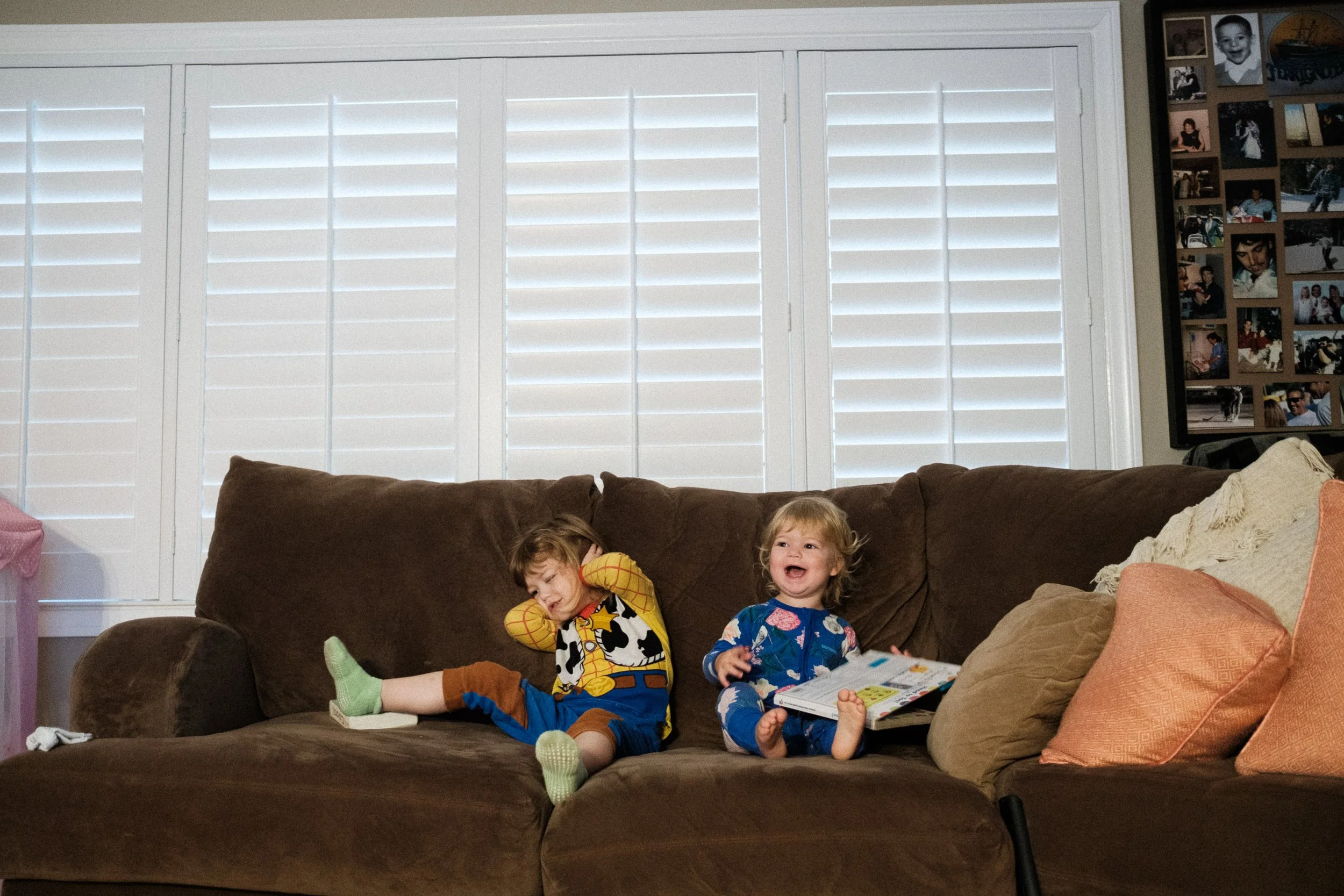 Two young children sitting on a brown sofa in a living room, one child appears distressed with hands on head, and the other is smiling and holding a book, with a framed photo collage on the wall behind them.