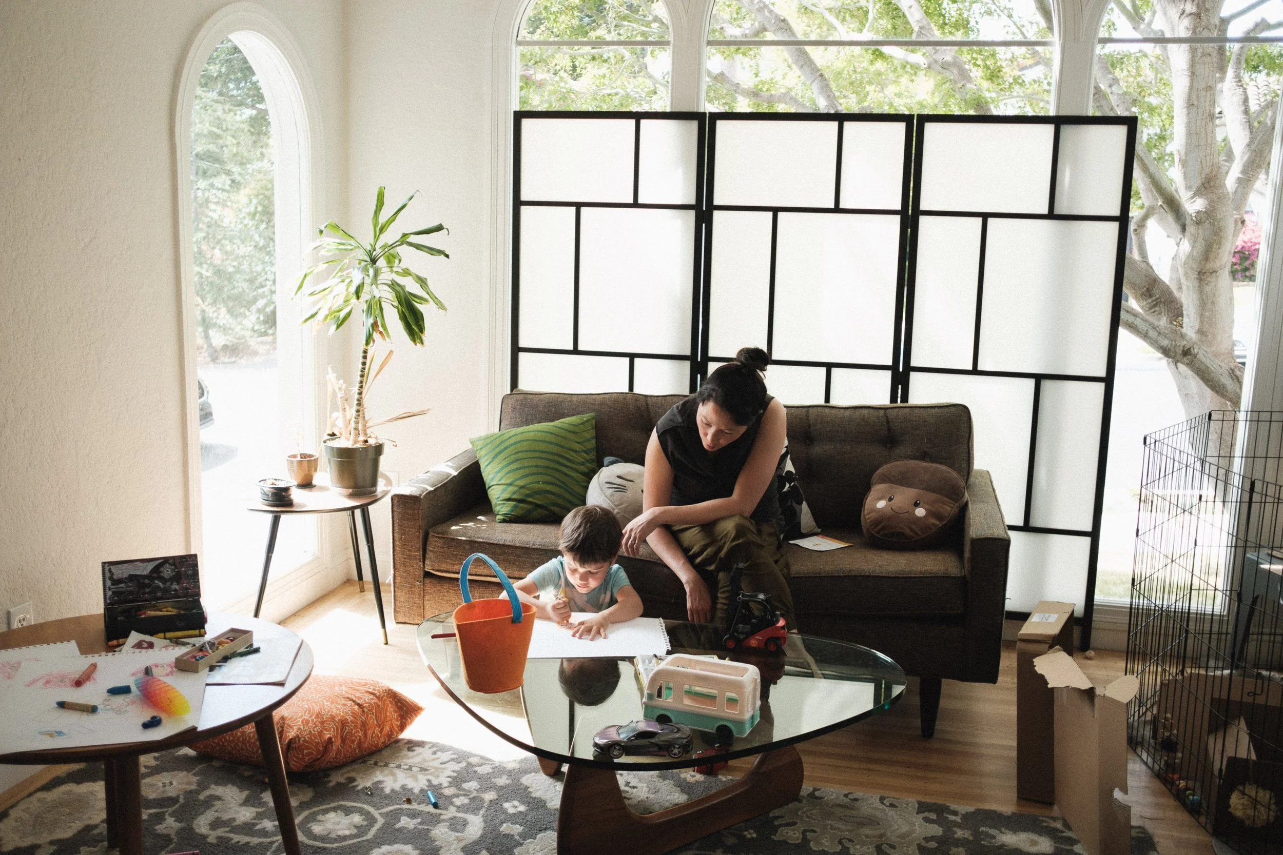 A woman and a young boy sitting on a couch in a living room. The boy is drawing at a glass coffee table with toys and a bucket. The woman is watching him. The room has large windows, a plant, and a folding room divider.