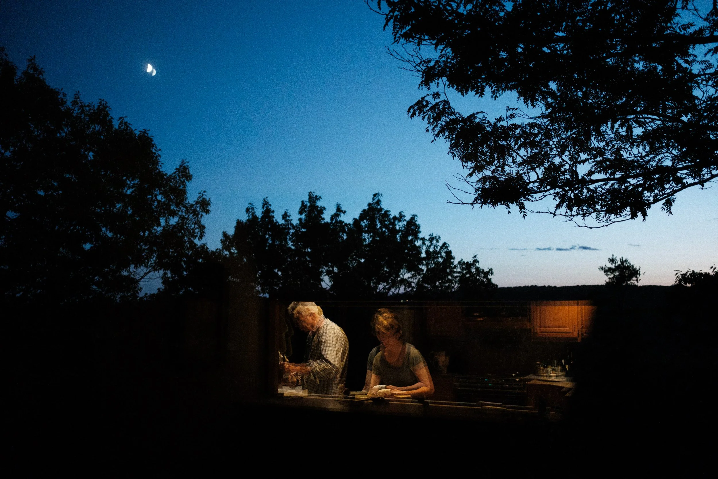 Two people cooking in a kitchen at night, with a dark outdoor scene and a crescent moon in the sky