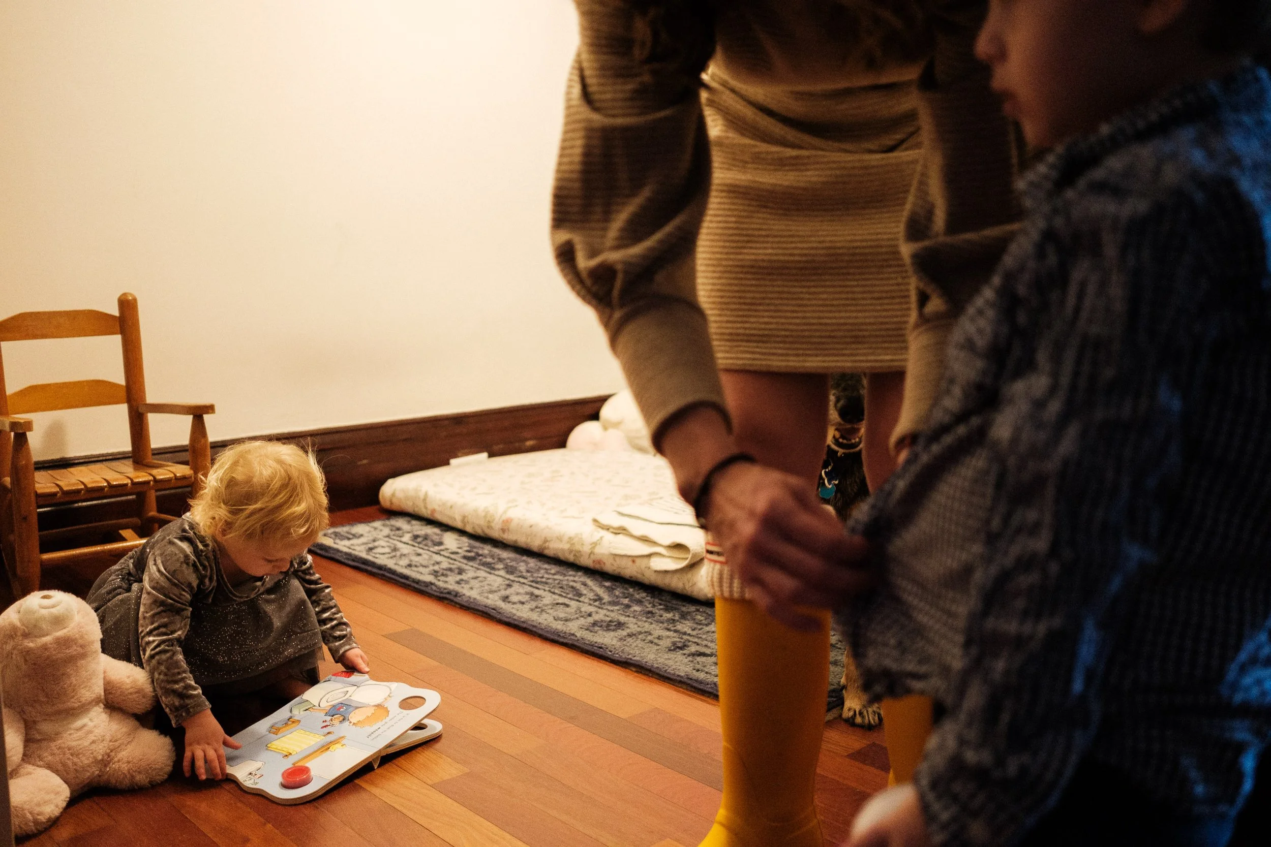 A young girl with blonde hair sitting on the floor next to a stuffed bear, looking at a picture book. A mother helps her son button his shirt. The scene is indoors with a bed and a chair in the background.
