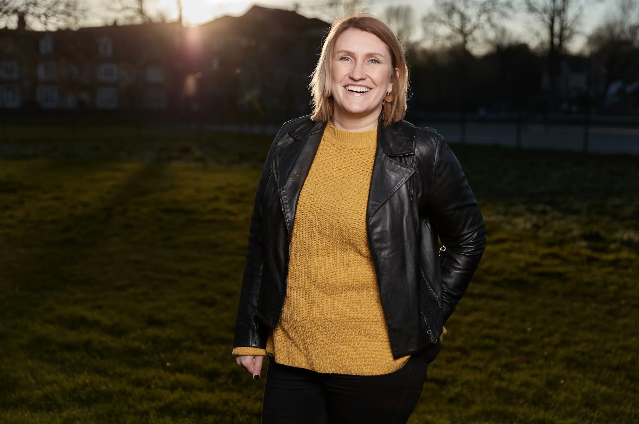 A woman smiling outdoors at sunset, wearing a black leather jacket over a yellow sweater, standing on grass with trees and houses in the background.