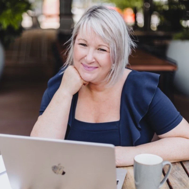 A woman with short gray hair sitting outdoors at a table with a laptop and a mug, smiling at the camera.
