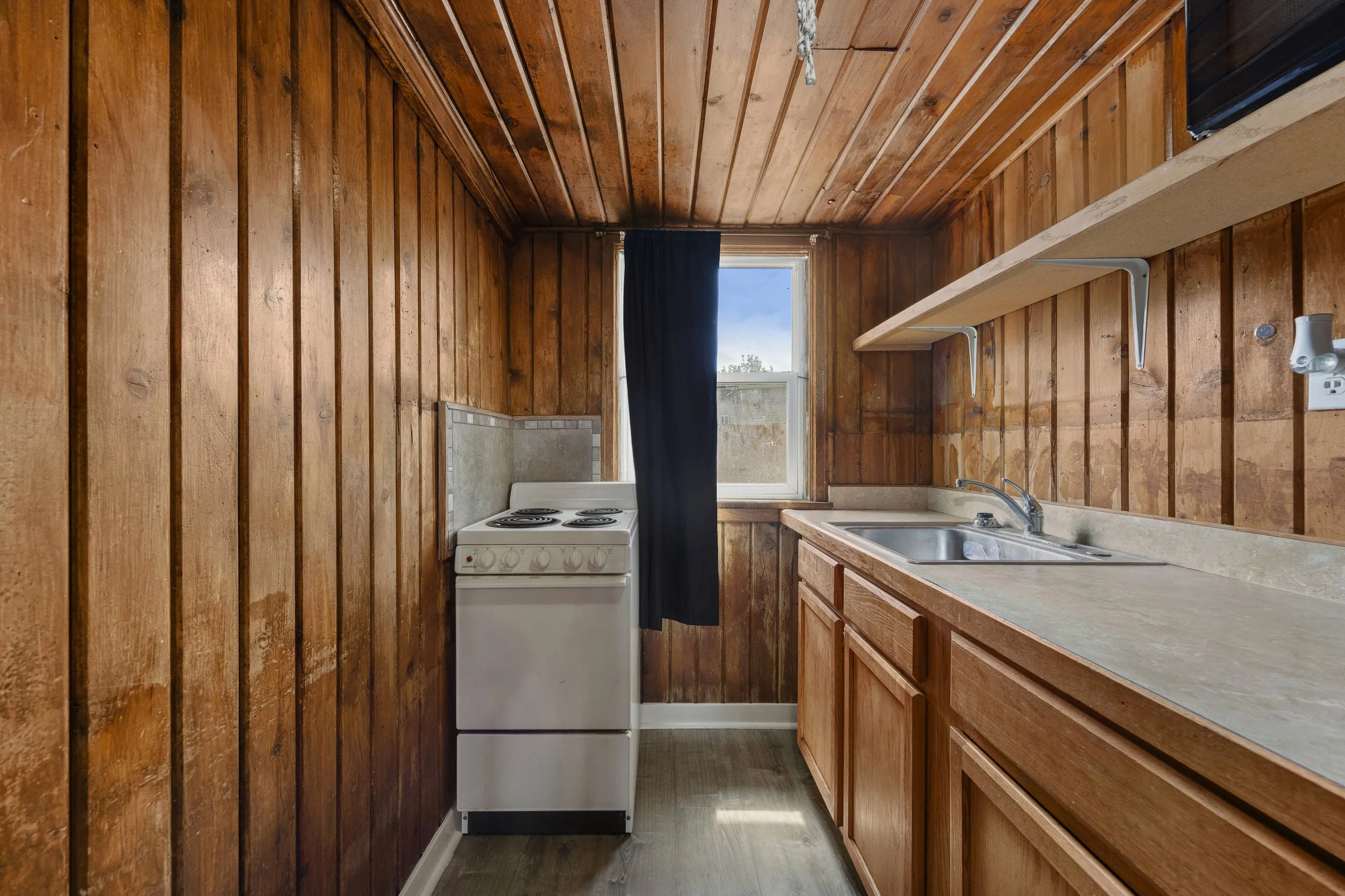 Small kitchen with wood-paneled walls and cabinets, a window with a black curtain, a white electric stove, and a sink with a faucet.