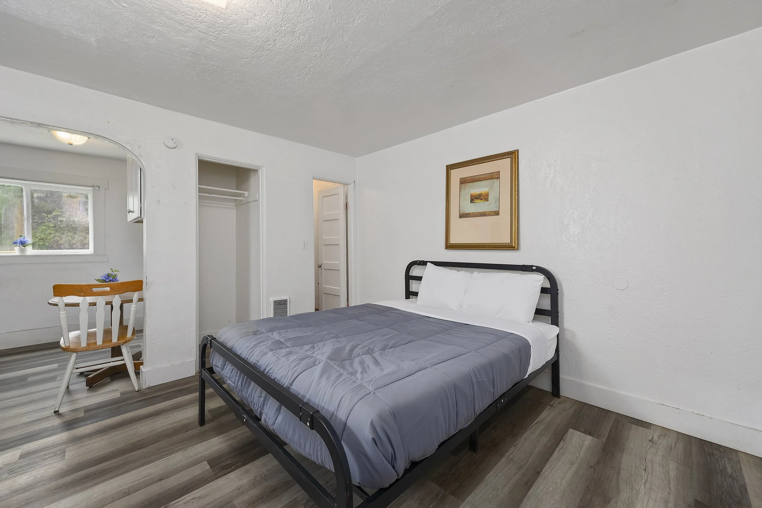 A bedroom with white walls and ceiling, a black metal bed frame with a gray bedspread, a painting on the wall, a window with a view outside, and a small dining table with a wooden chair.