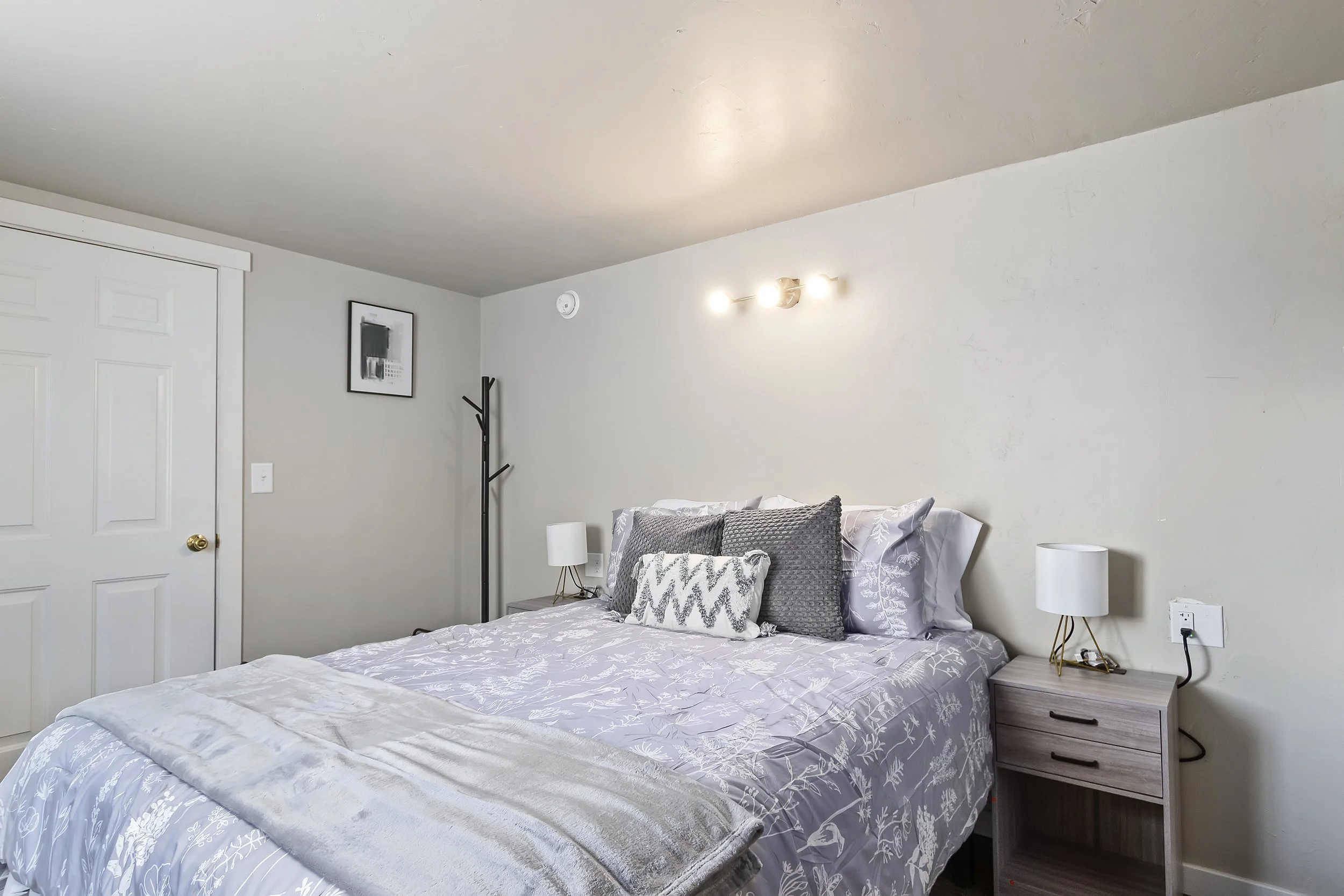 A bedroom with a bed covered in a light-colored floral comforter, gray pillows, two white nightstands with lamps, a black coat rack, a framed picture on the wall, a closed white closet door, and a ceiling light fixture with three bulbs.