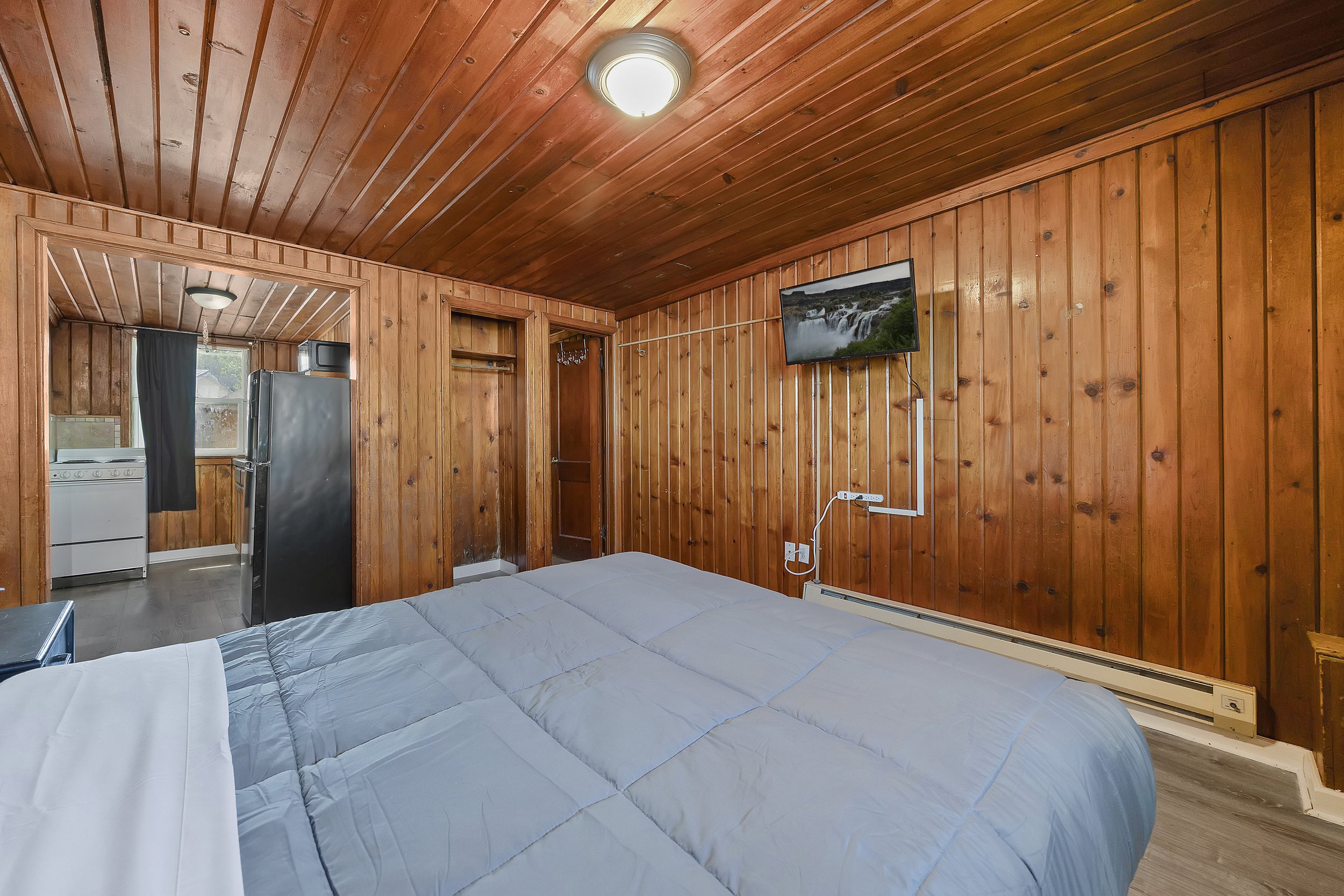 A cozy wooden room with a bed covered in light gray bedding, a wall-mounted TV showing a waterfall scene, and a view into a small kitchen area with a refrigerator and stove.