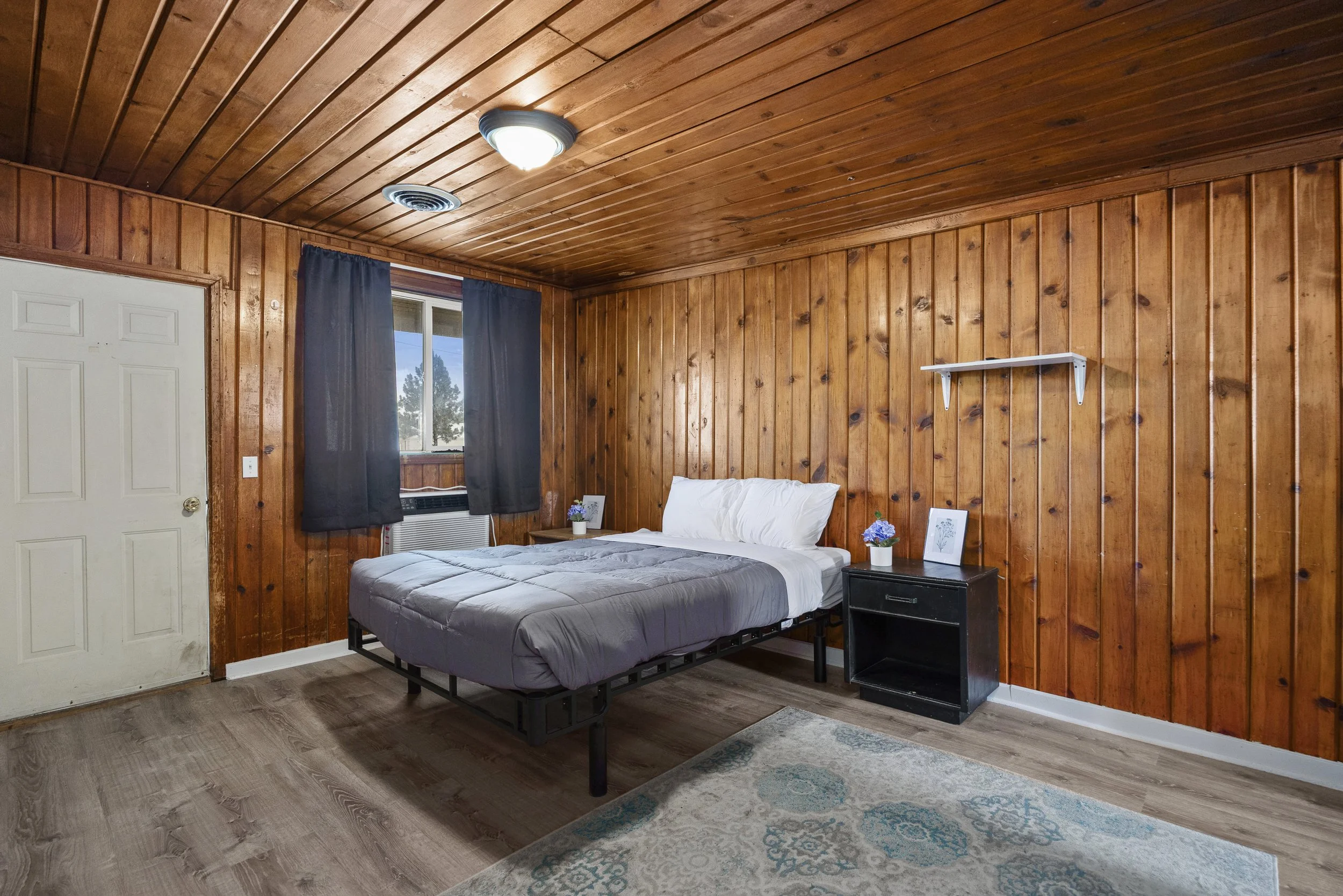 A bedroom with wood-paneled walls and ceiling, a window with dark curtains, a bed with white bedding, a black nightstand with potted flowers, and a white shelf on the wall.