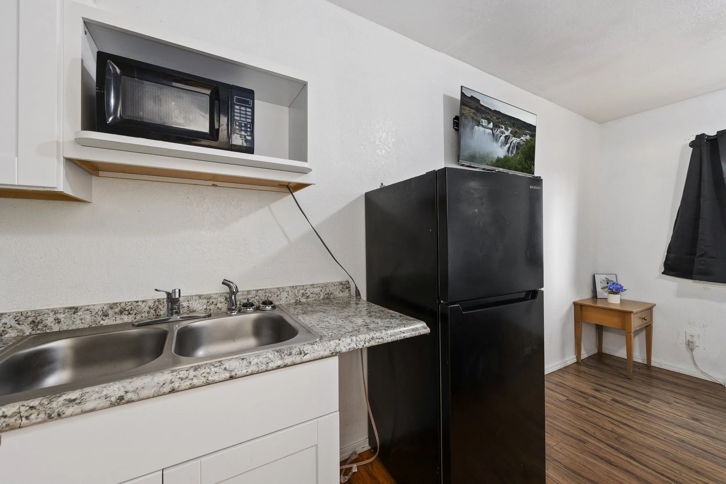 A kitchen with a granite countertop, double sink, microwave in a wall niche, black refrigerator, and a small wooden table with a potted plant.