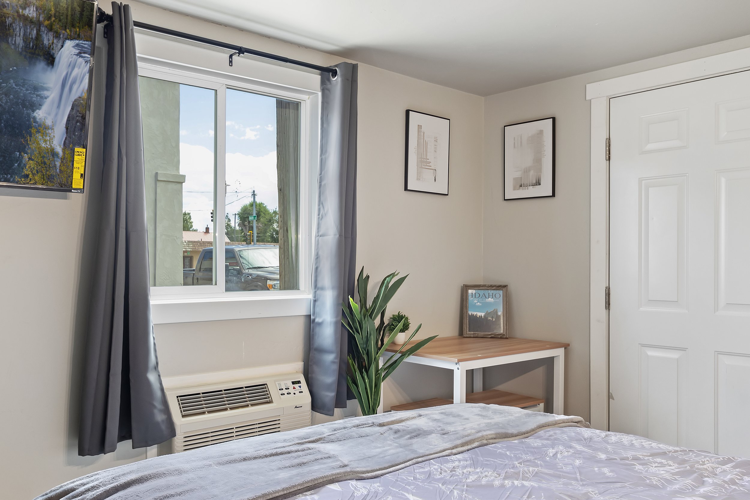 A bedroom with a large window showing a street scene, curtains, framed artwork, a potted plant, and an air conditioning unit below the window.