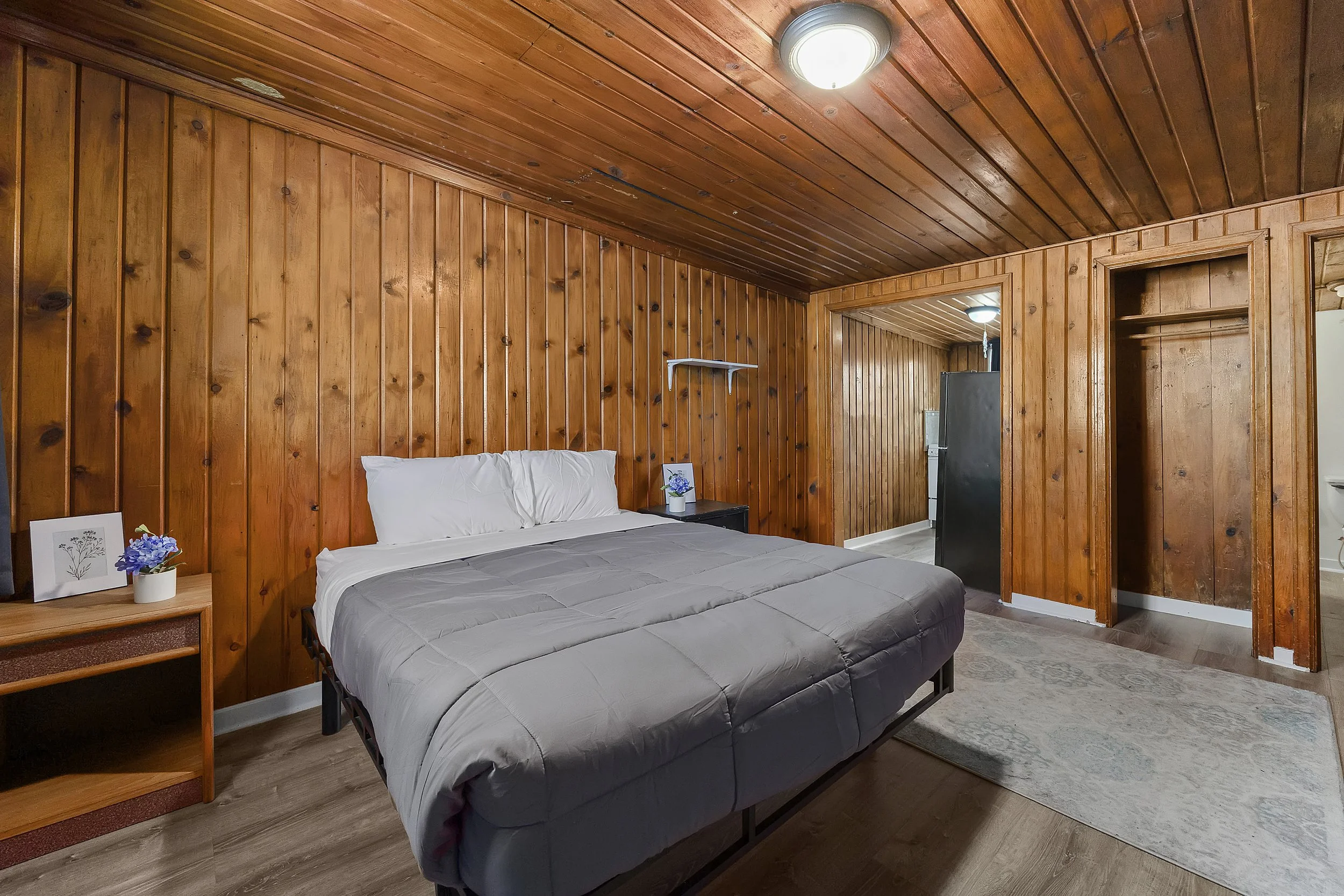 A bedroom with wood-paneled walls and ceiling, a bed with gray bedding, nightstand with a vase of purple flowers, and an open doorway leading to another room.