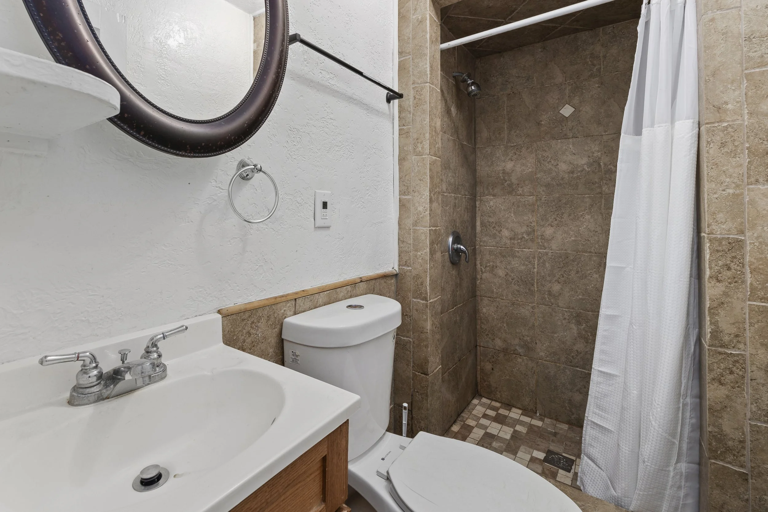 A small bathroom with a white sink, faucet, and wooden vanity, a toilet, and an empty shower area with beige tiled walls and a white shower curtain.