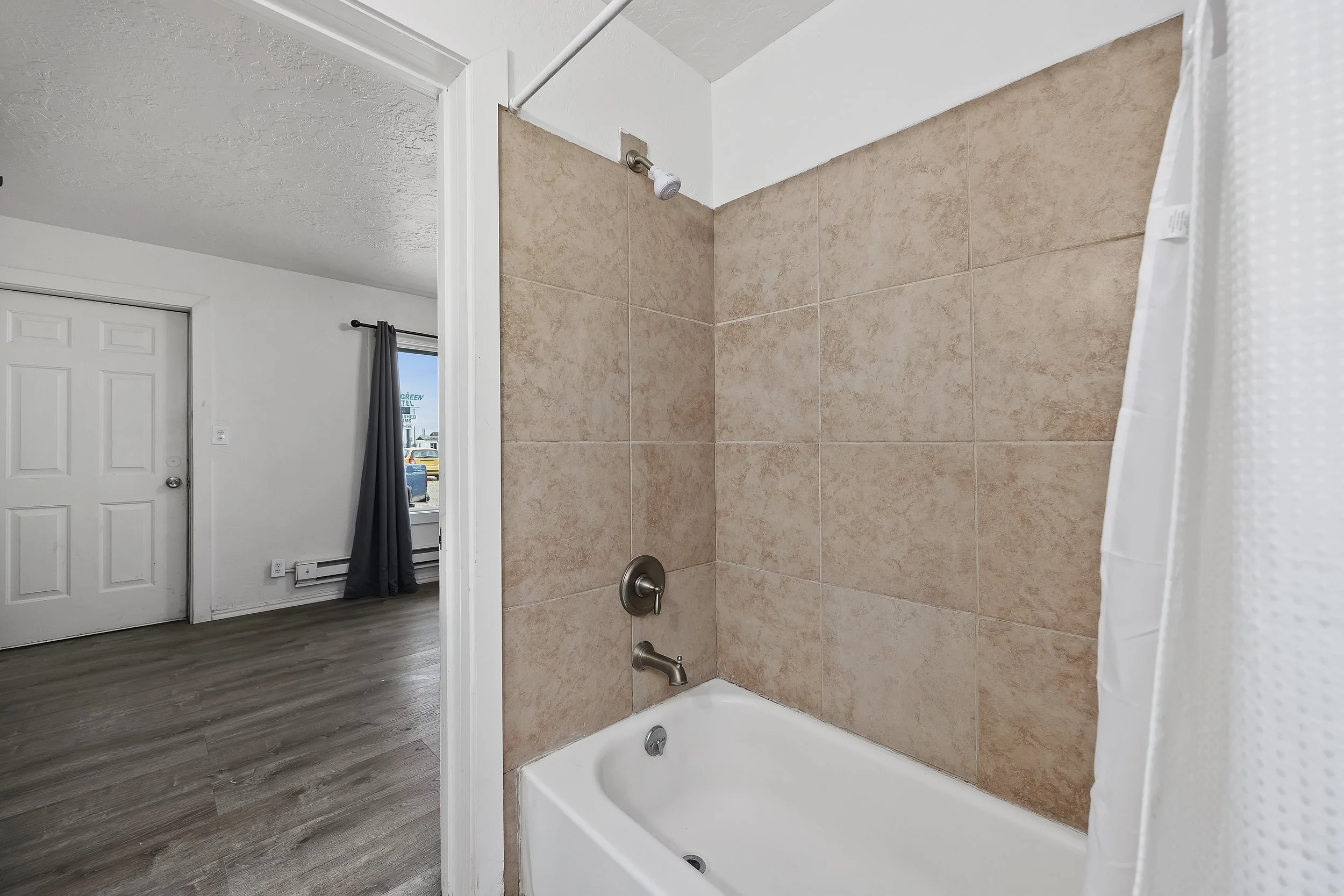 A bathroom shower with beige tiled walls, a white bathtub, a showerhead mounted on the wall, and a partially visible shower curtain. The bathroom has white walls and a door in the background, with a window showing a parking lot outside.
