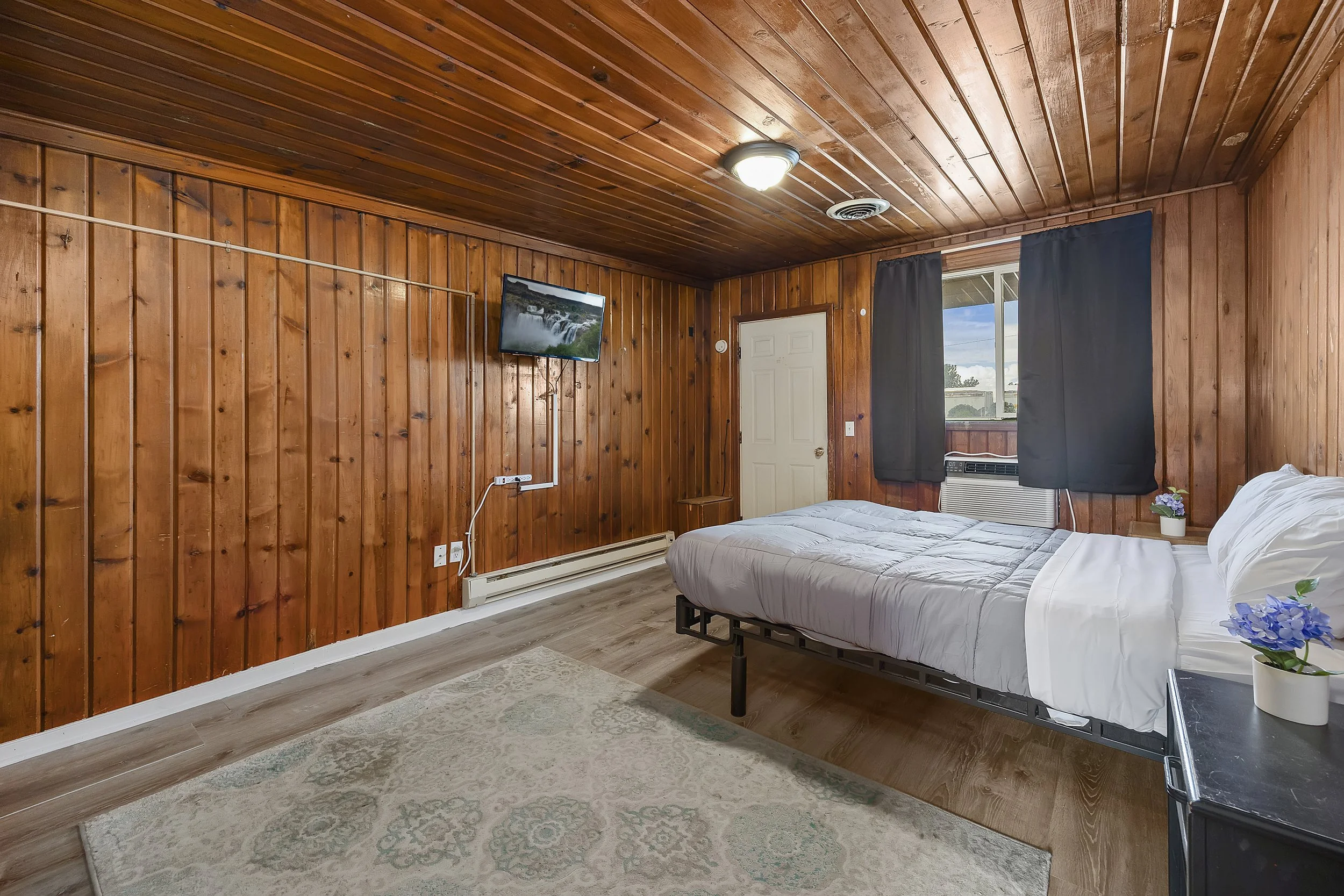 A bedroom with wood-paneled walls and ceiling, a bed with white linens, a window with black curtains, a wall-mounted TV, and a small table with a potted plant.