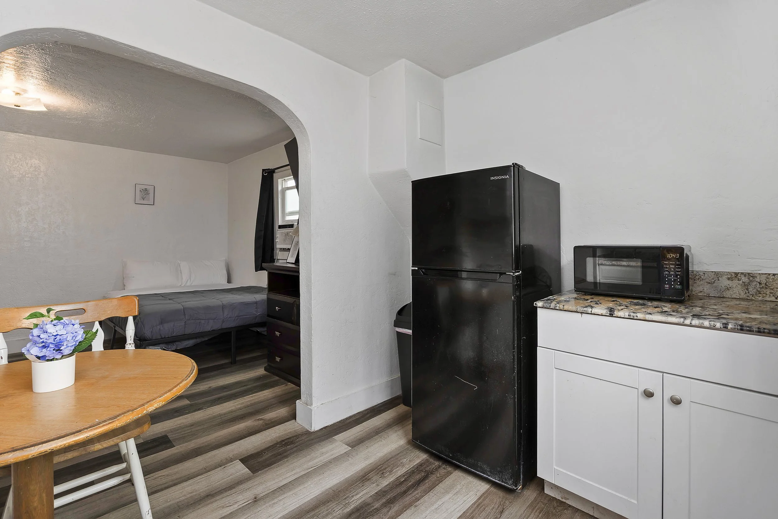 Interior view of a kitchen with a black refrigerator, microwave, white cabinets, and a granite countertop. A small dining table with a floral centerpiece, white chairs, a bed in the background with a picture on the wall, and a window with dark curtai