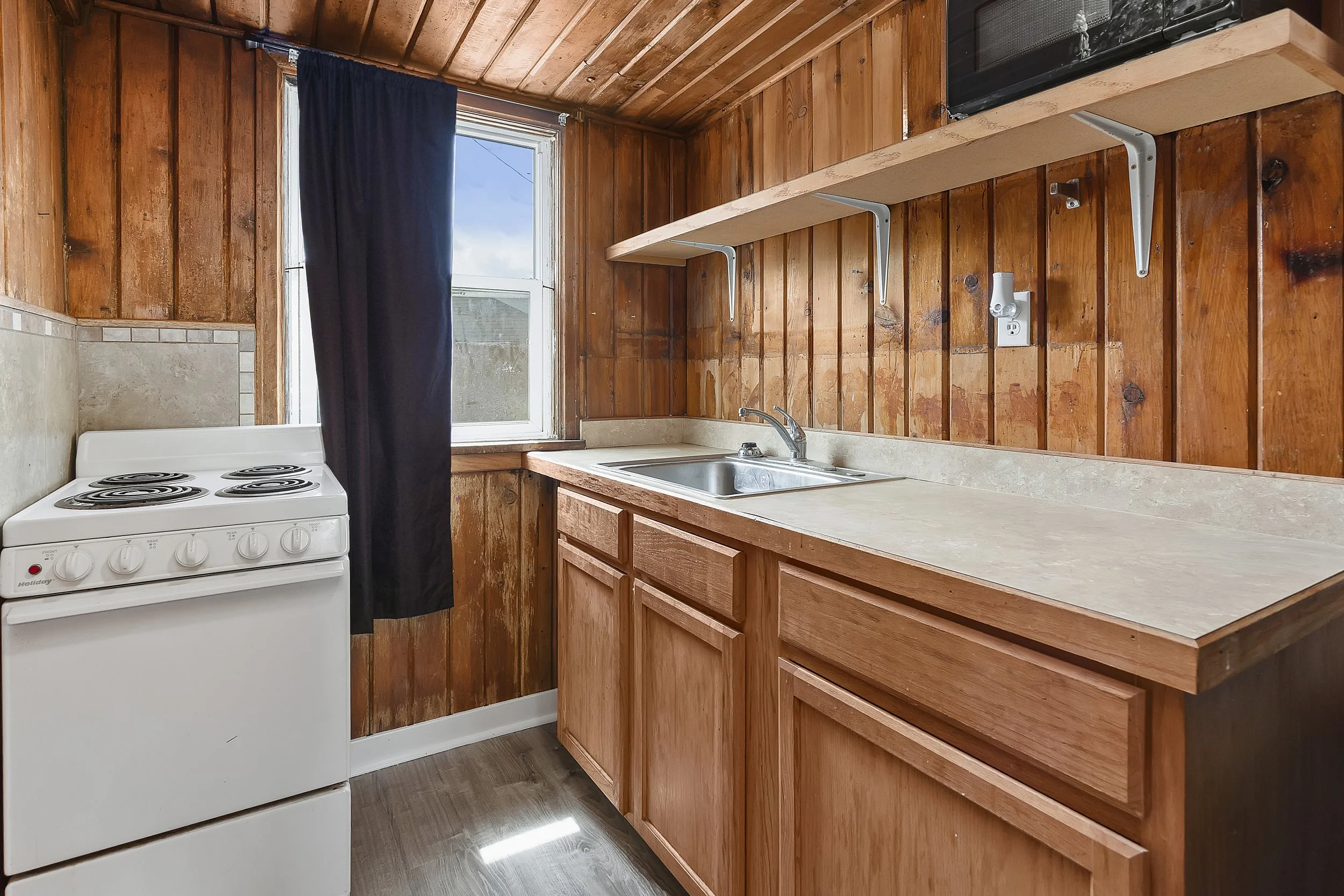A small kitchen with wood-paneled walls and ceiling, a white stove on the left, a countertop with a sink and faucet, a window with a dark curtain, and a black microwave on an open shelf above the counter.