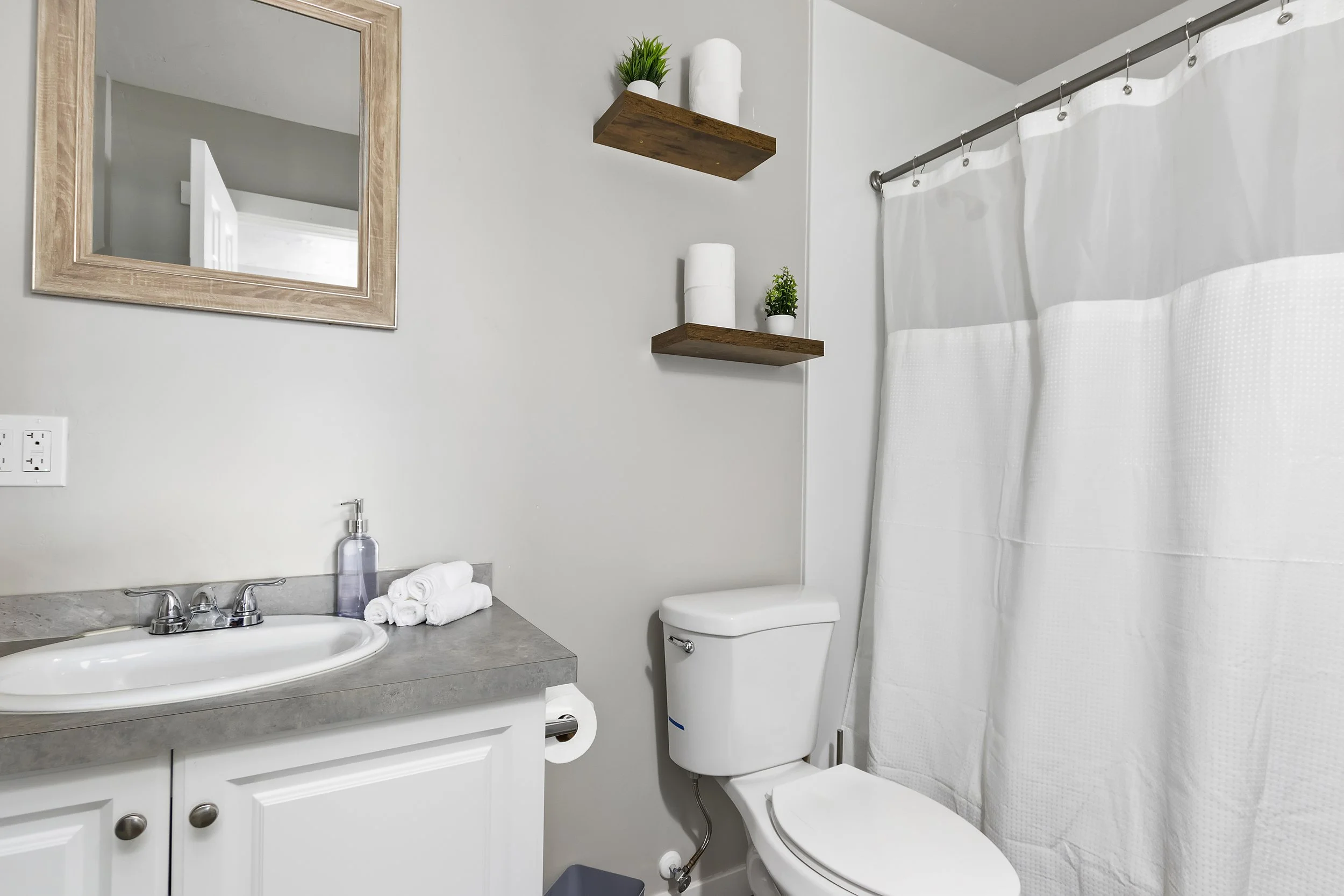 A bathroom featuring a white countertop with a sink, a faucet, and a soap dispenser. Above the sink is a rectangular mirror with a wooden frame. To the right of the mirror, there are two wooden shelves holding small potted plants and decorative items