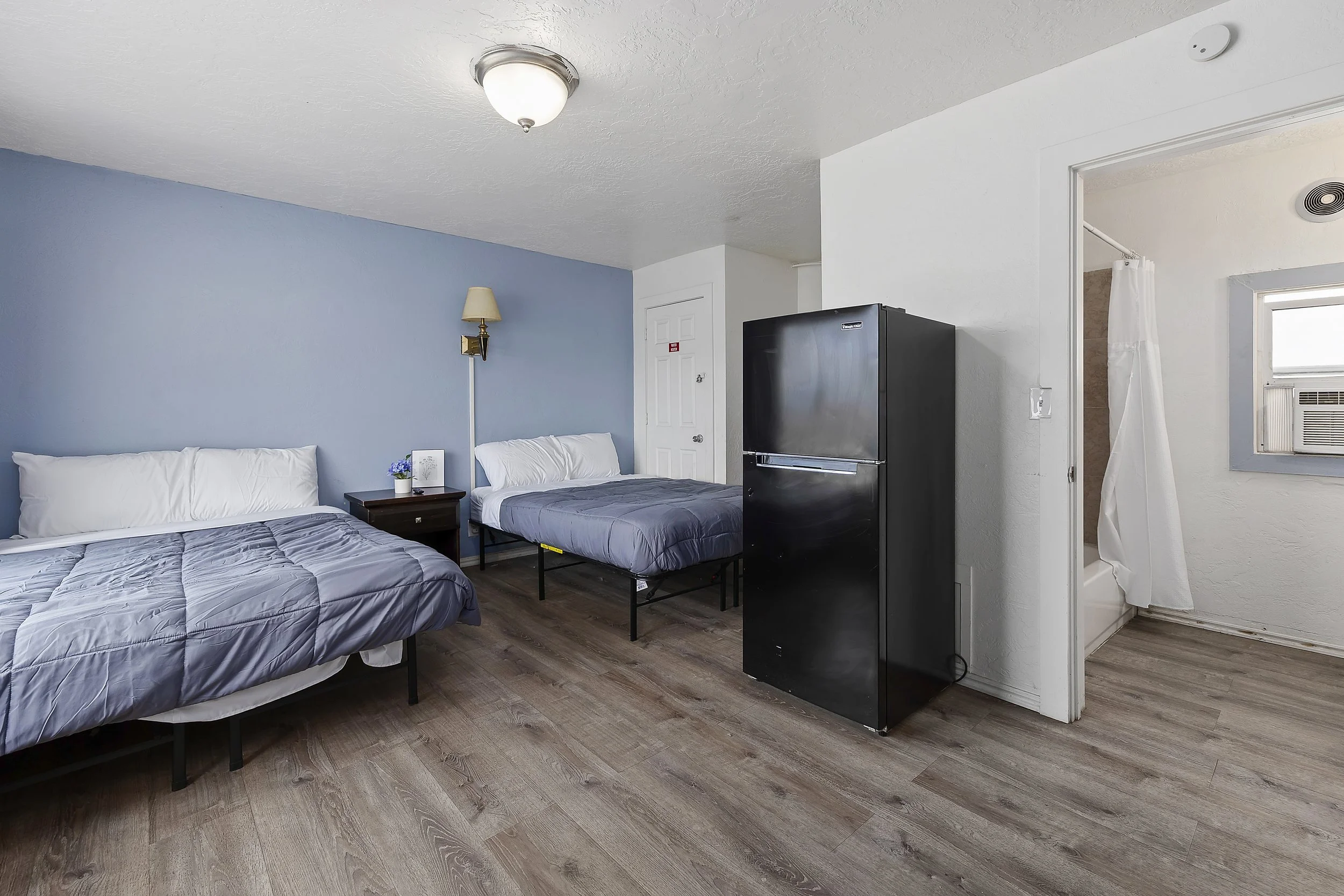 Room with two beds, blue and white walls, a nightstand with a small flower pot, a wall-mounted lamp, a refrigerator, and a doorway leading to a bathroom with a window and an air conditioning unit.