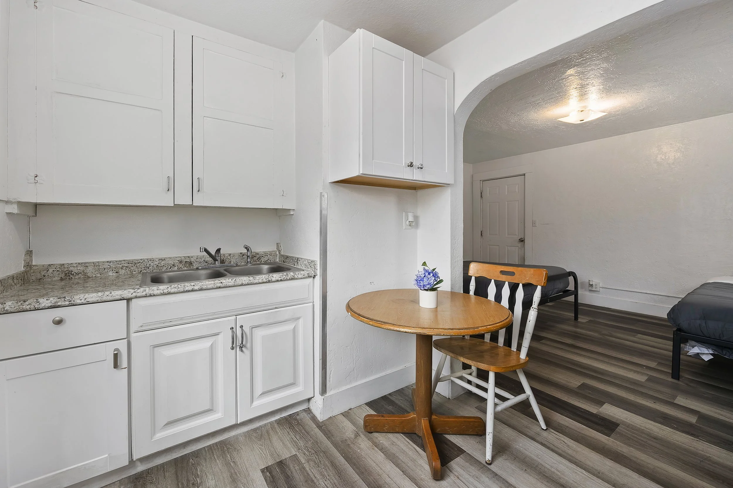 A small kitchen with white cabinets, a granite countertop, and a twin sink. Adjacent is a dining area with a round wooden table, a white chair, and a plant in a white pot. In the background, there is a dark coffee table and a bed, with a door and a c