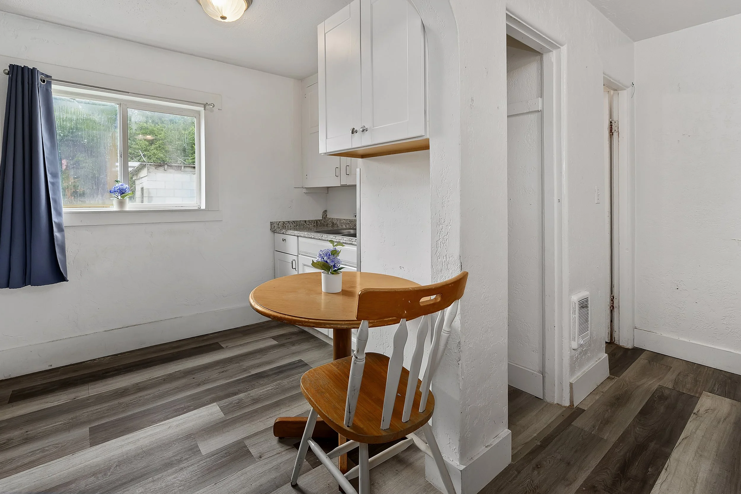 A small kitchen with a round wooden dining table and two wooden chairs with white frames. A potted plant with purple flowers is on the table. There is a window with a blue curtain, and outside trees are visible. The kitchen has white cabinets, a gran